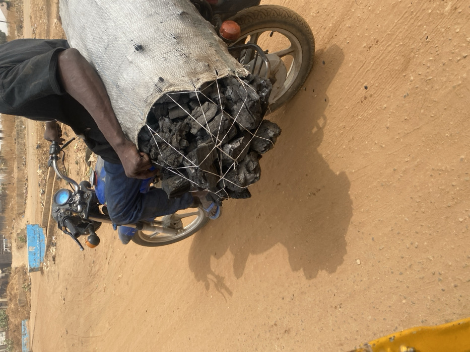 A man rides a motorcycle laden with a large sack of charcoal. The charcoal is bound by white netting, overflowing from the bag onto the motorcycle's front. The rider wears dark clothing and blue sandals, his dark skin contrasting with the bright tan dirt road. The bike's shadow stretches across the ground, suggesting a sunny day.