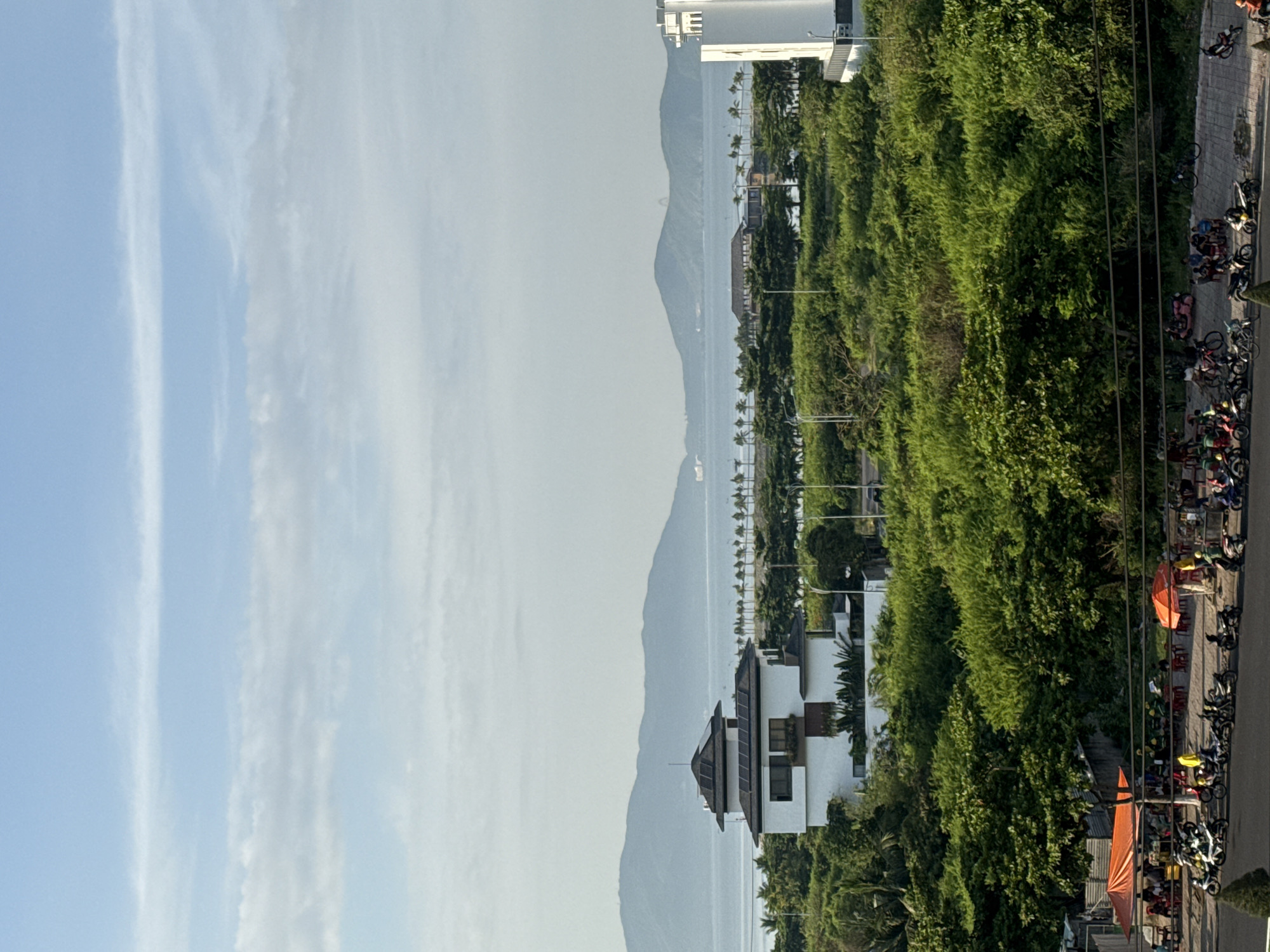 The image shows a landscape scene in Bac Nha Trang Ward, Vietnam. In the foreground, a street is lined with parked motorcycles and people sitting under orange umbrellas, possibly at a market or roadside eatery. Beyond the street, a dense area of lush green trees stretches across the middle ground. Prominent structures are visible within the greenery, including a white building with a dark, tiered roof and solar panels, and a tall, white modern building on the right. In the background, a body of water separates the land from a range of hazy, green mountains under a pale blue sky with scattered clouds. A ship is visible on the water in the far distance. The overall impression is a coastal view with a mix of natural landscape and urban development.