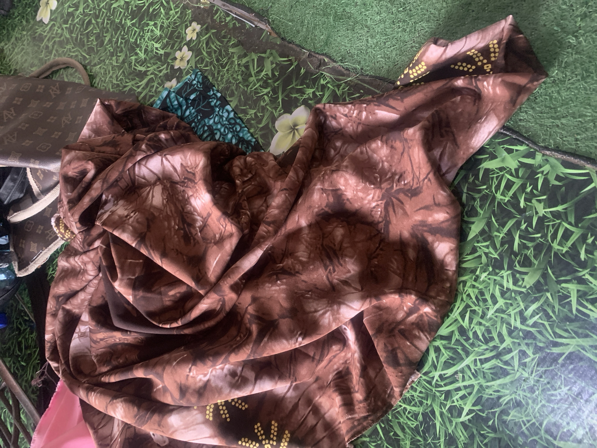 A close-up, overhead shot shows a brown, patterned fabric draped over a green, artificial grass mat. The fabric has a tie-dye-like pattern in shades of brown and black, with some areas appearing slightly iridescent. On the left side of the fabric, a section with small, yellow dots is visible. In the background, a portion of a brown designer handbag with a repeating logo pattern and a strap is visible, along with a pink piece of fabric and some dark objects that appear to be branches or sticks. The mat has a few small, white flowers scattered across it. The scene appears to be indoors, possibly in a home environment in Jauro Yinu, Nigeria.