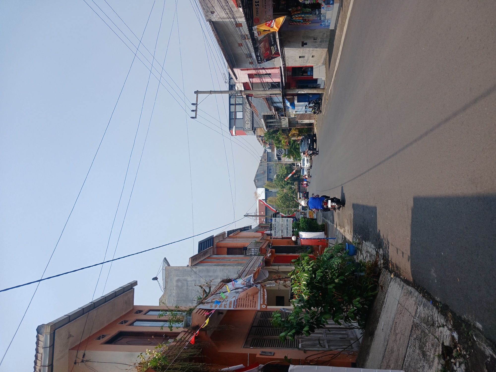 The image depicts a street scene in Watugede, Indonesia, under a clear, bright sky. The perspective is from a low angle, looking down a paved road lined with buildings on both sides. The buildings are a mix of two and three-story structures with a variety of architectural styles and colors, including orange, pink, and gray. Many of the buildings appear to be commercial establishments, with signs and displays visible. On the left side of the road, a motorcycle rider in a blue jacket and helmet is traveling away from the camera. Several other vehicles, including cars and motorcycles, are also visible further down the road. Trees and lush green foliage are interspersed with the buildings, adding a touch of nature to the urban environment. Electrical wires are strung across the sky, crisscrossing the scene. The road surface is asphalt, and a sidewalk runs along the side of the buildings. Shadows from the buildings and trees are cast on the road, indicating the time of day is likely late morning or early afternoon. The overall impression is one of a bustling, everyday street in a developing urban area.
