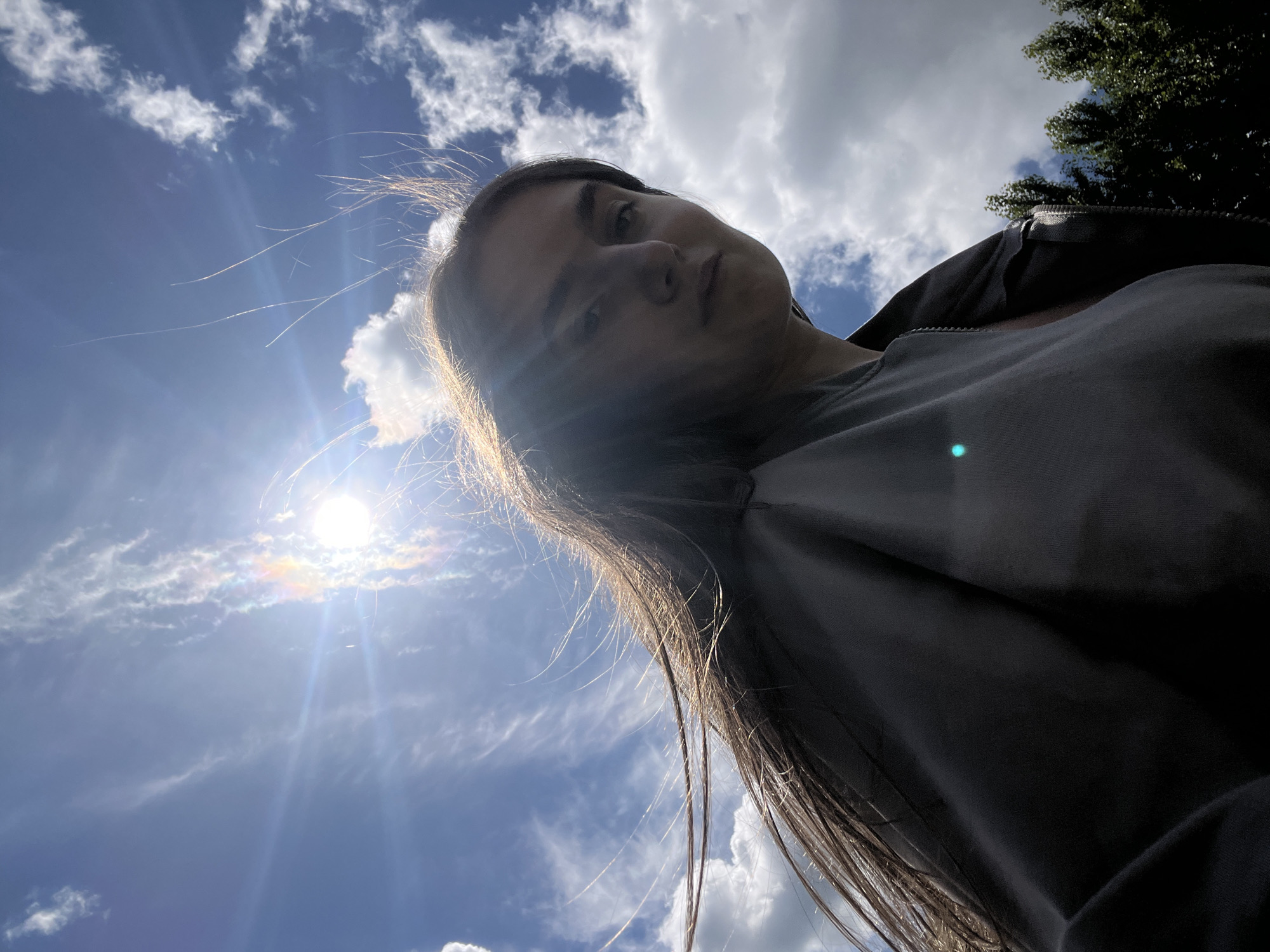This is a low-angle, close-up, outdoor selfie taken in Ternopil, Ukraine. The image features a young woman with long, dark hair blowing in the wind. She is wearing a dark-colored jacket and is looking towards the camera with a neutral expression. The sun is bright and high in the sky, creating lens flares and illuminating the clouds. The sky is a vibrant blue with scattered white clouds. In the background, the tops of green trees are visible. The overall lighting suggests it is a sunny day.