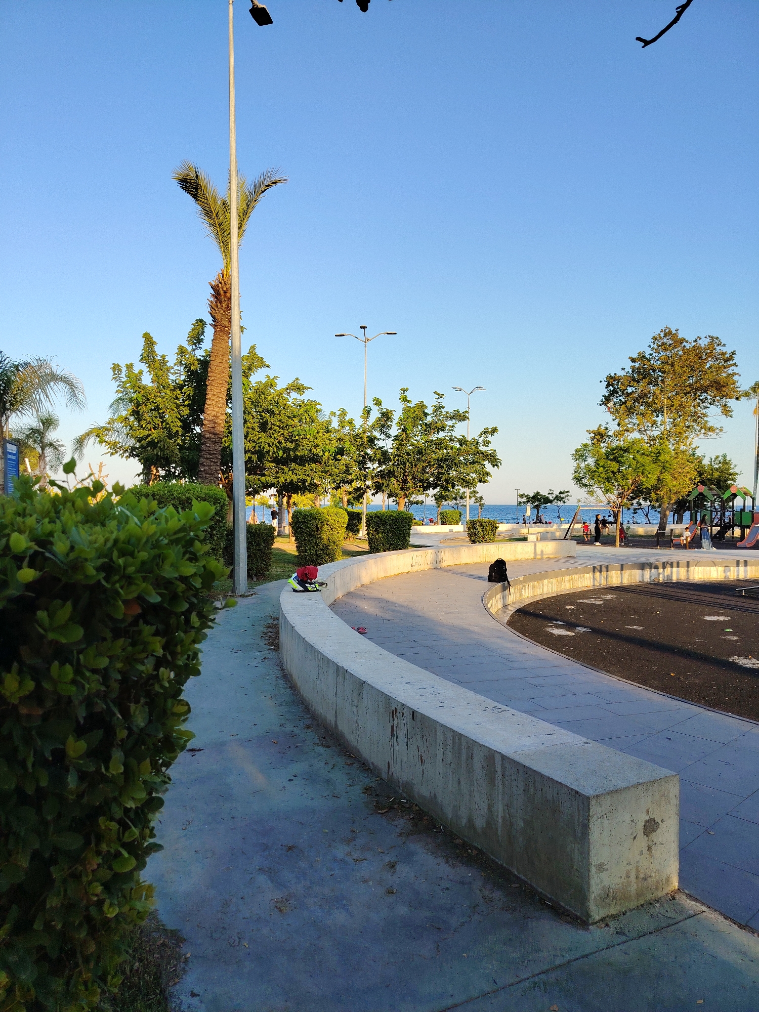 The image shows a paved public area with a curved concrete wall in the foreground. To the left, a dense green hedge partially obscures a concrete walkway. Behind the wall, a dark asphalt surface is visible, possibly part of a playground or recreational area, with some scattered leaves. In the middle ground, a palm tree stands tall next to other leafy green trees. A lamppost is situated between the palm tree and the hedge. Further back, the sea is visible with people walking along the shore and a playground with colorful structures. The sky is clear and blue. The scene suggests a public park or promenade in a coastal city, likely in a warm climate. Based on the vegetation and architecture, this scene is consistent with the coastal areas of Antalya, Turkey, possibly in a park like the one along Konyaaltı Beach or near the Old Town (Kaleiçi) with its sea views.