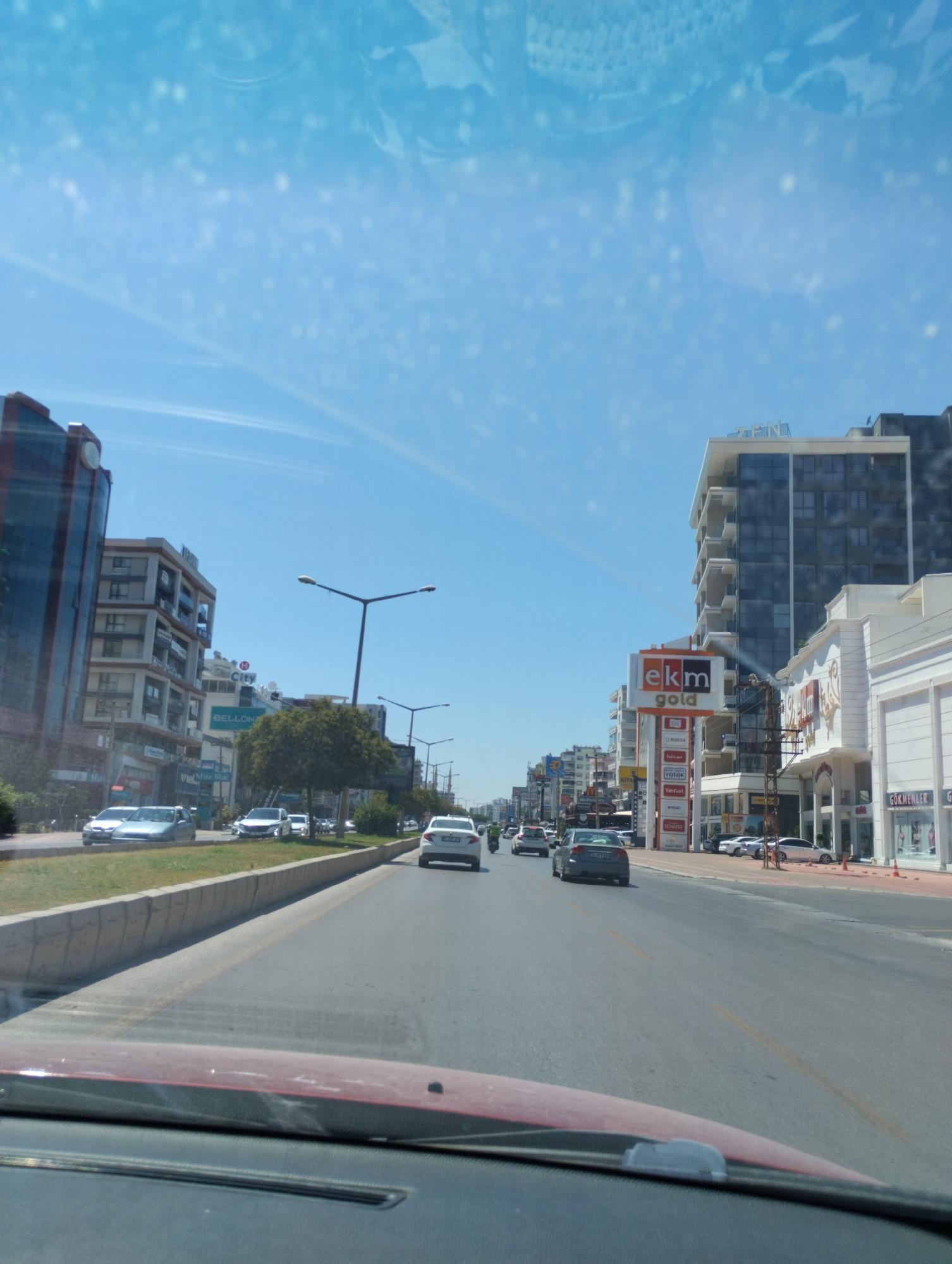 The image depicts a street scene in Mezitli, Turkey, likely during daylight hours under a clear blue sky with scattered white specks, possibly from the camera lens or a light haze. The perspective is from inside a vehicle, looking forward. A multi-lane road dominates the foreground, with cars traveling in the same direction. On the right side of the road, a row of modern buildings is visible. One prominent building features a large sign for "ekm gold" and also displays logos for various brands like Bosch, Tefal, and Schafer. Further along the right, another building has signage indicating "GÖKMENLER". On the left side of the road, a taller building with a dark facade and circular clock is visible, followed by several other multi-story structures. Trees are interspersed along the median and sidewalk areas. Streetlights are present on both sides of the road. The overall impression is of a commercial and residential area within a city.