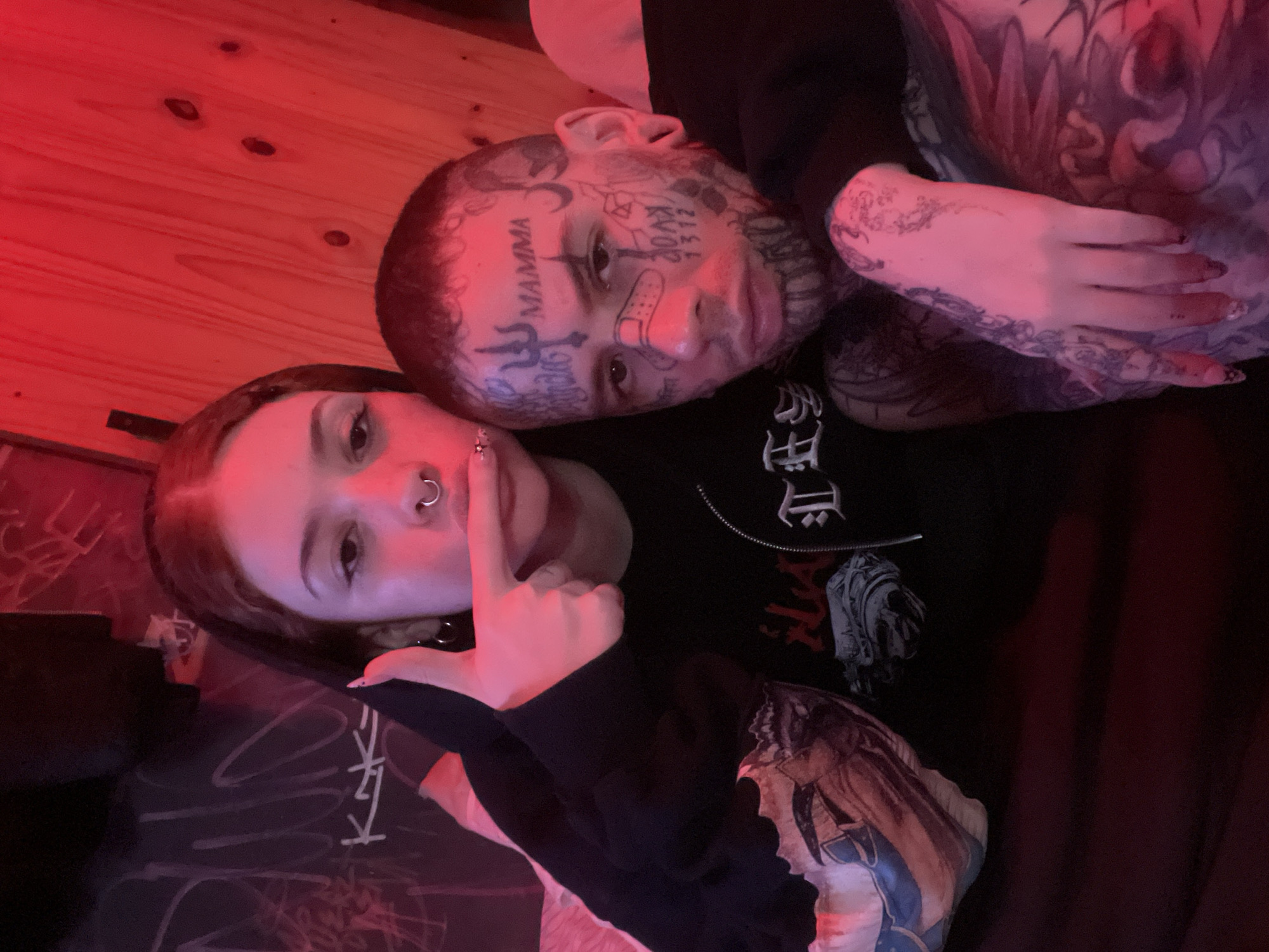Two people are lying next to each other. The person on the left is a light-skinned young woman with reddish-brown hair, a nose ring, and a black choker necklace. She is holding a finger up to her lips. The person on the right is a light-skinned individual with numerous face tattoos, including writing on their forehead and symbols on their cheeks. They also have a bandage on their nose. Both individuals are wearing dark-colored clothing, and the clothing of the person on the right features white writing and a graphic design. The scene is illuminated with red light. A wooden surface appears in the background. The location is not apparent, and it is unclear if the image was taken in Buenos Aires, Argentina.
