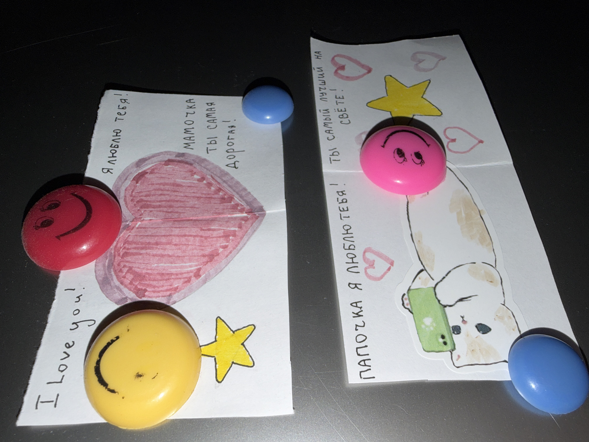 The image shows two pieces of paper on a dark, reflective surface, likely a refrigerator door. The left paper features a drawing of a pink heart, a yellow star, and three round smiley face magnets (red, yellow, and blue). Text in English "I Love you!" and in Russian "Я люблю тебя! МАМОЧКА ТЫ САМАЯ Дорогая!" (Mommy you are the dearest!). The right paper features a drawing of a cat taking a selfie, two hand-drawn pink hearts, a yellow star, and three round magnets (pink, blue, and blue). Russian text reads "ΠΑΠΟΣΚΑ Я ЛЮБЛЮ ТЕБЯ! Ты Самый лучший на СВЁТЕ!" (Daddy I love you! You are the best in the world!). The text, images, and magnets suggest these are handmade notes or drawings for parents, attached to the refrigerator. The location is unknown.