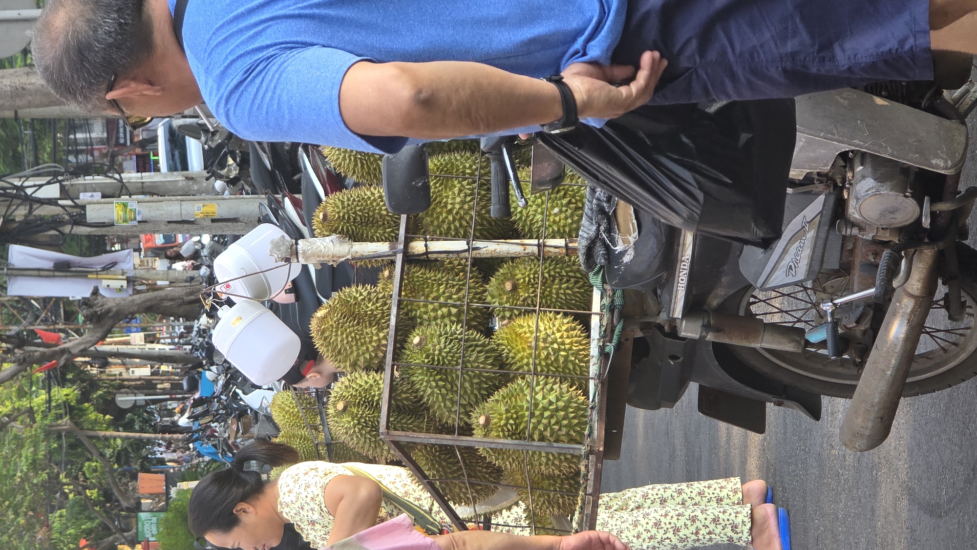 A street scene shows two people near a motorbike loaded with durian fruits. A man in a blue shirt and dark pants stands near the motorbike, examining the fruit. On the other side, a woman in a floral top and pants looks at the durians. The motorbike has a metal cage attached, containing numerous durians. Two white buckets hang near the front of the motorbike. The motorbike, labeled "HONDA Dream II", is parked on the roadside. In the background, a busy street includes more parked motorbikes, utility poles with wires, and trees. The setting is Nha Trang, Vietnam.