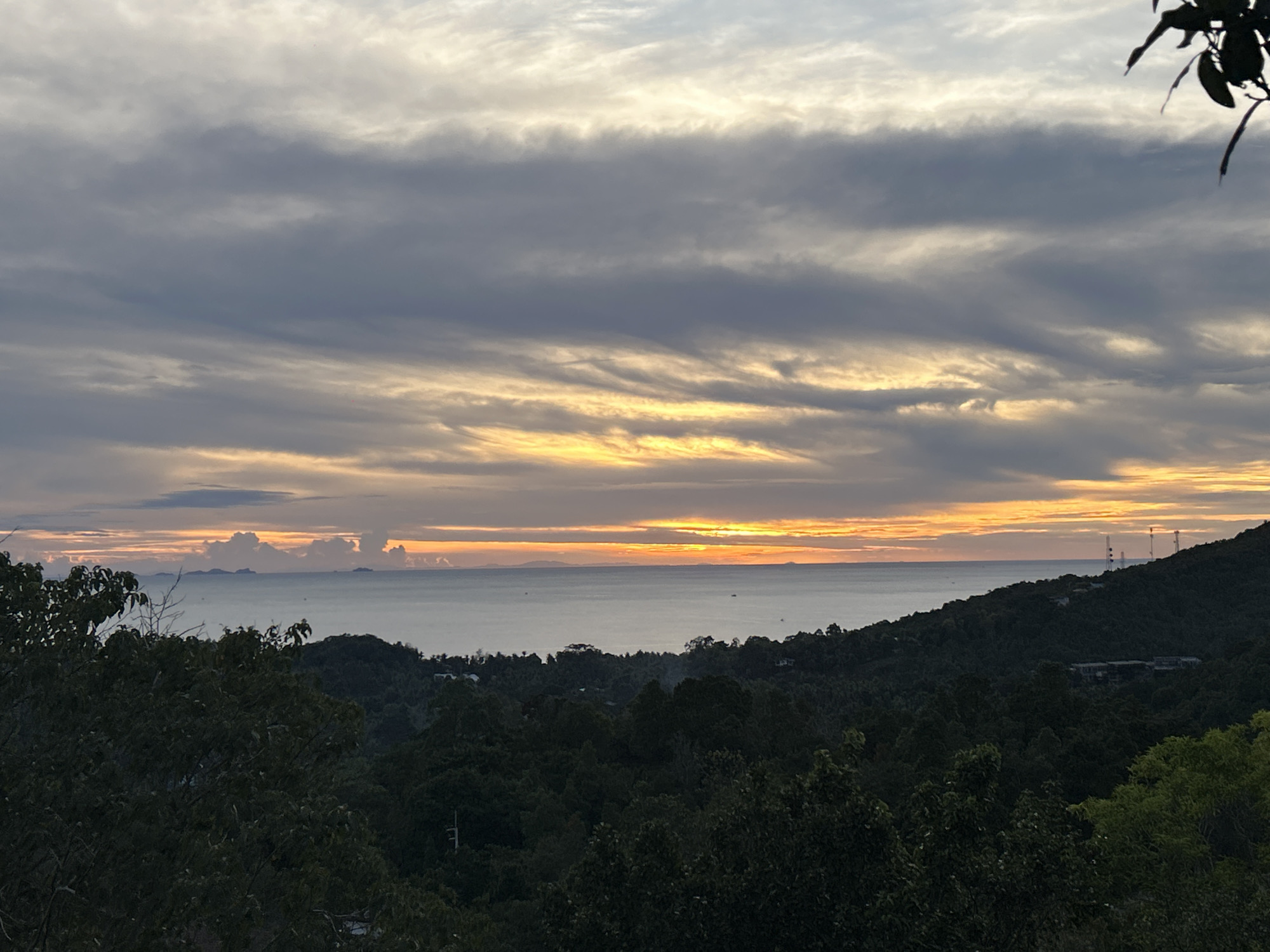 The image shows a sunset over the sea, likely taken from an elevated position in the Baan Hin Kong area of Thailand. The sky is filled with layers of clouds, colored with hues of grey, yellow, and orange from the setting sun. Below the horizon is the calm sea, with some islands visible in the distance on the left. The foreground is dominated by lush, dark green vegetation, suggesting a dense forest or jungle environment. On the right are the trees and some buildings with tall cellular towers.