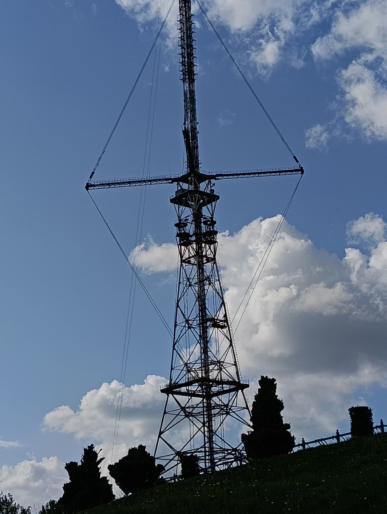 The image captures a silhouette of a tall, lattice metal tower against a bright blue sky dotted with white clouds. The tower, with its intricate network of beams and supports, dominates the frame, extending upwards towards the sky. Several guy wires extend from the tower, anchored to the ground at various points, creating a web-like structure. At the base of the tower, the ground slopes upwards, covered in grass and silhouetted trees. A dark, low fence is visible along the crest of the hill. The overall scene suggests an outdoor, open space, possibly a park or elevated area within the city. The tower is likely a telecommunications tower or a similar structure. Without further context or identifying markers within the image, its exact location within Hrodna, Belarus, cannot be definitively determined from the visual information alone.