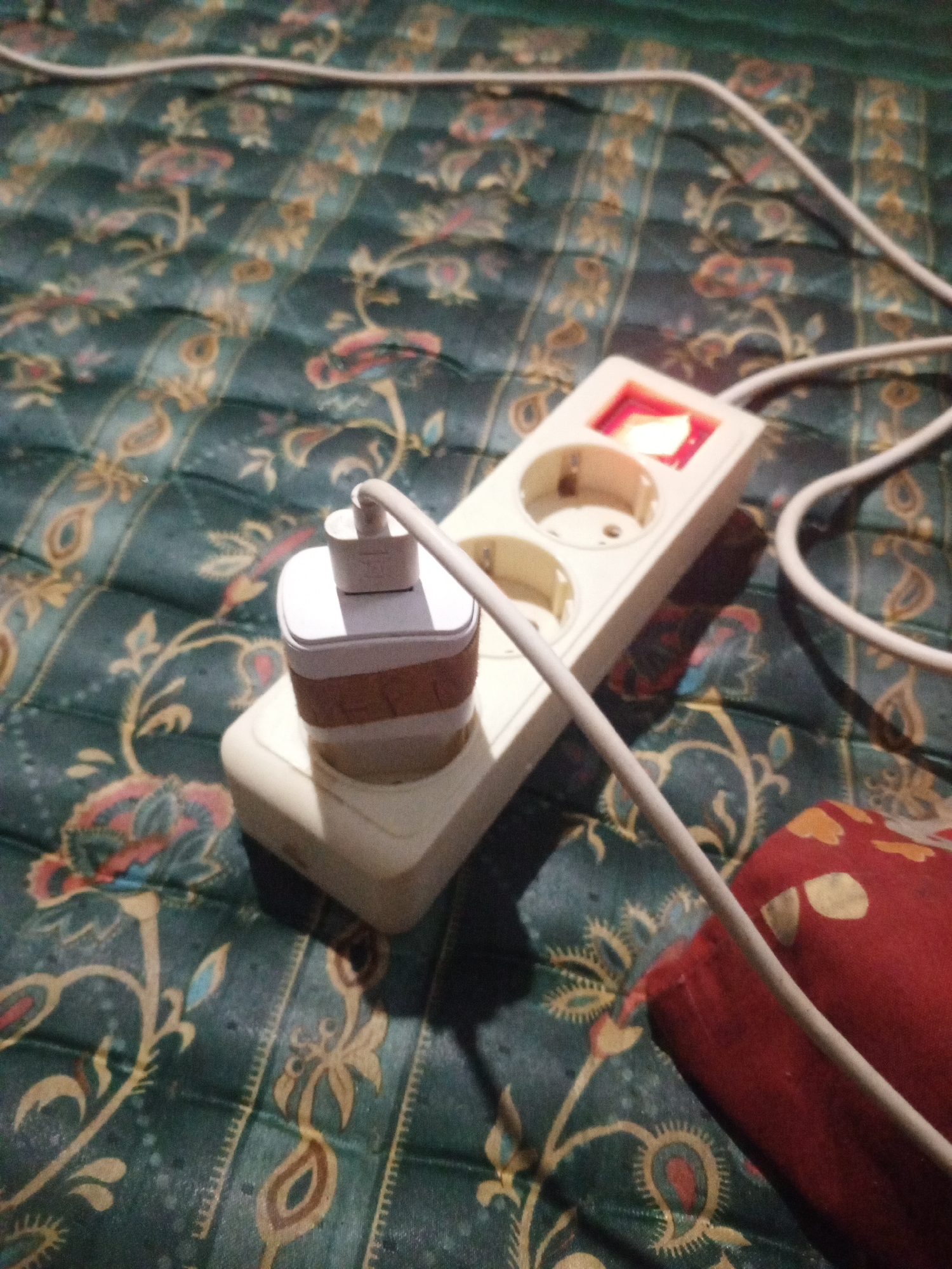 In Tanah Gara Hulu, Indonesia, a white power strip with two outlets is plugged into a patterned surface, possibly a mattress or comforter with a dark green base and an intricate floral design in shades of red, yellow, and blue. A white phone charger adapter is plugged into one of the outlets, and a white cable extends from the adapter across the surface. A red object, possibly a pillow or cushion, is partially visible to the right of the power strip. The scene appears to be indoors, likely in a residential setting, given the domestic context of a power strip and charging adapter.