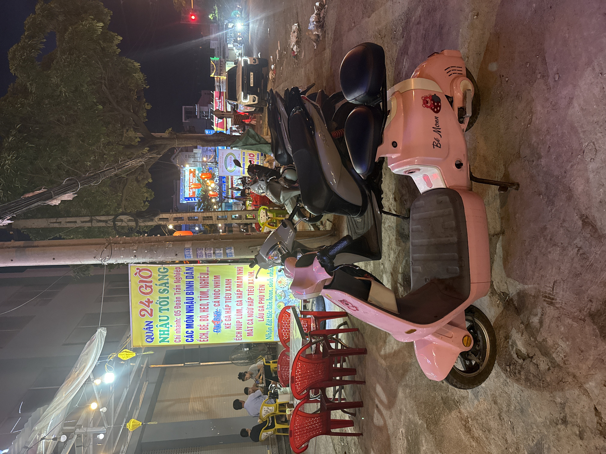 The image depicts a street scene at night in Nha Trang Ward, Vietnam, likely outside a restaurant or food establishment. A bright yellow banner hangs on a building to the left, advertising "QUÁN 24 GIỜ NHẬU TỐI SANG" which translates to "24-Hour Restaurant for Evening Gathering." The banner lists various dishes, including "CÁC MÓN NHẬU BÌNH DÂN" (common bar snacks), "ẾCH, BÈ, BÒ, HEO, TÔM, NGHỀU..." (frog, shellfish, beef, pork, shrimp, clam...), with specific mentions of "Đặc Biệt: CÁ NÓC NHÍM" (Special: Pufferfish sashimi), "KÈ GÀ HẤP TIỂU XANH" (Chicken and vegetable steamed dish), and "ẾCH NÚP LÙM, GÀ HẤP MĂM NHÍ" (Hidden frog, seasoned steamed chicken).
In the foreground, a pink electric scooter is parked on the sidewalk, with a "Bé Monn" logo and a cartoon bear sticker visible on its side. The scooter has a dark grey seat and handlebars. Behind the scooter, several other motorbikes are parked, including a grey one and a darker one with a prominent red rear brake light.
To the left of the scooter, four red plastic chairs surround a table, indicating an outdoor seating area for the establishment. A group of four people, two adults and two children, are seated at another table further back. They appear to be dining. A large industrial fan is visible behind them.
The background shows a tree with dense foliage, illuminated by streetlights and signs. A white pickup truck with license plate "90A-370.78" is parked on the street to the right, with its headlights on. The street is lined with other businesses, indicated by illuminated signs. The ground is a concrete sidewalk and street, littered with debris in some areas. A traffic light is visible in the upper right corner, showing a red light. The overall atmosphere suggests a lively street scene at night.