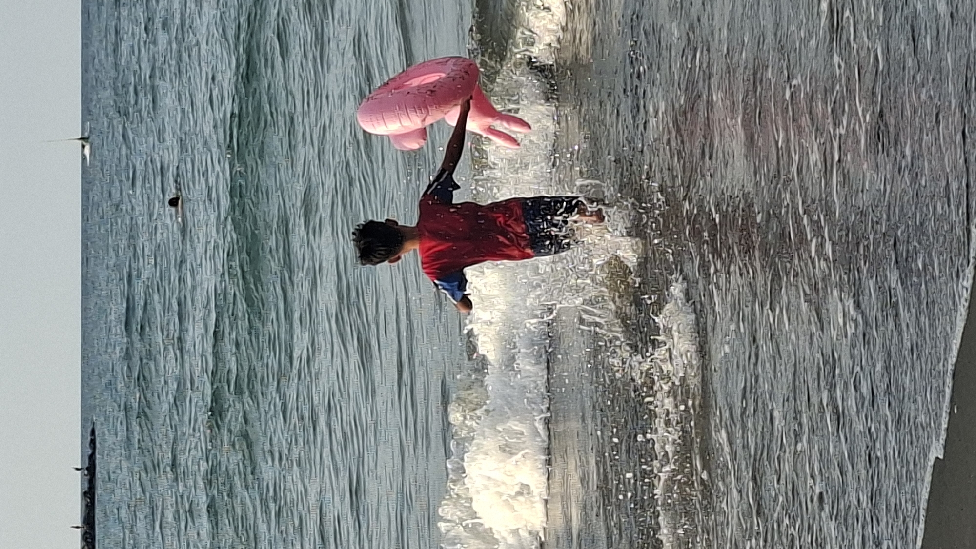The image depicts a young boy running into the ocean at a beach, holding a pink inflatable toy. He is wearing a red t-shirt with blue sleeves and dark shorts, and he is kicking up water as he runs. Waves are crashing behind him, creating white foam. In the distance, there are a few other people in the water, and a small boat or sailboard is visible. The sky is overcast, and the water appears somewhat choppy. The overall scene is one of a child enjoying a day at the beach. This scene could be occurring at a beach in Lhokseumawe, Indonesia.