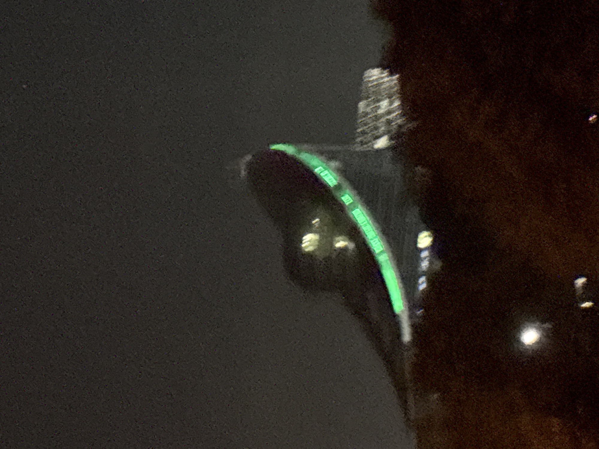 The image shows a partial view of the Sofia Eye Ferris wheel in Sofia, Bulgaria at night. The Ferris wheel has a green-lit structure. In the background, a blurred building is visible. The foreground on the right is dark and textured.