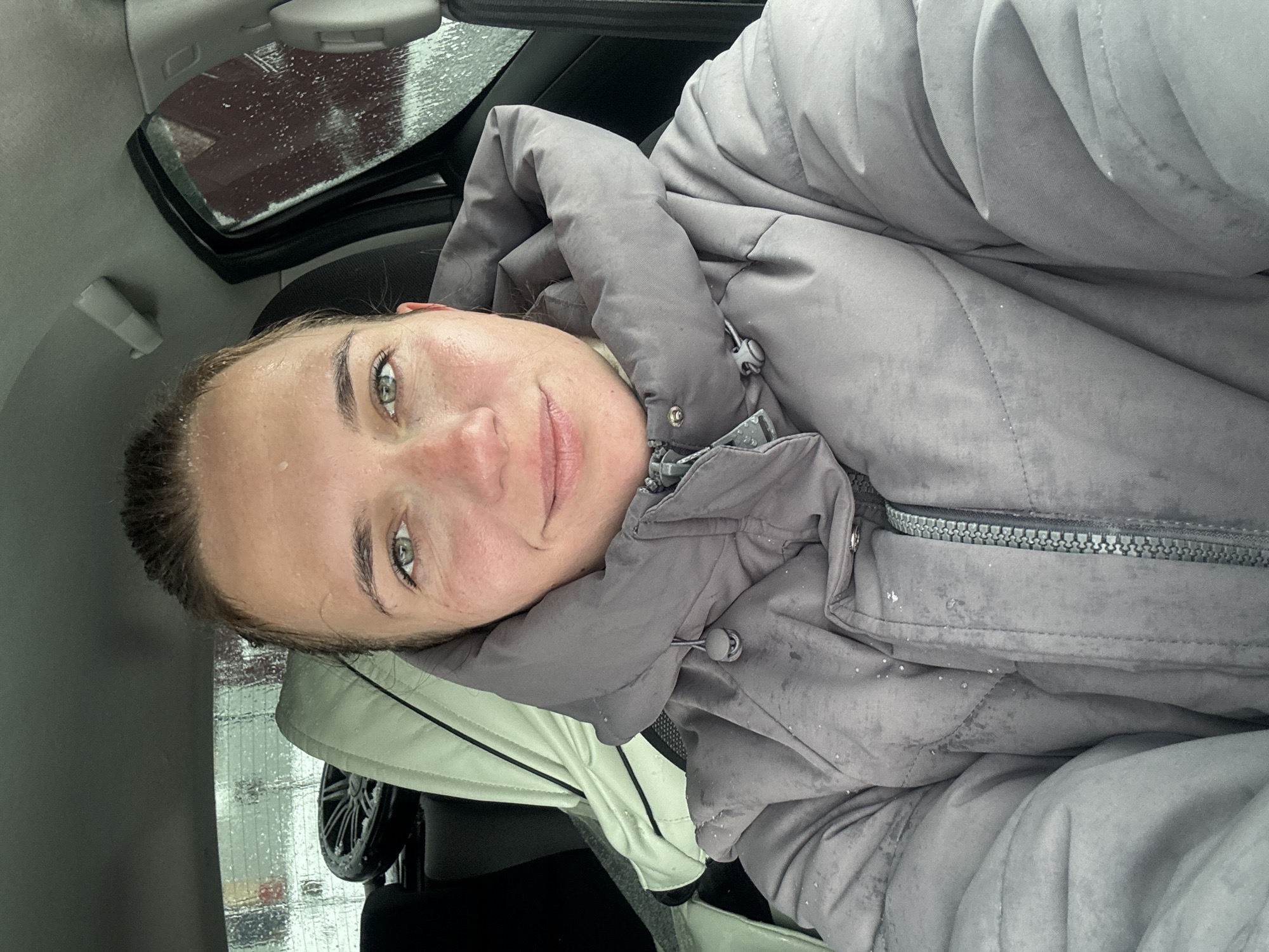 The image shows a woman in a gray puffer jacket, likely sitting in a car. Raindrops are visible on the car windows. The location is possibly Раздолье, Russia, though this is based on the prompt, and there is no confirming information in the image itself.