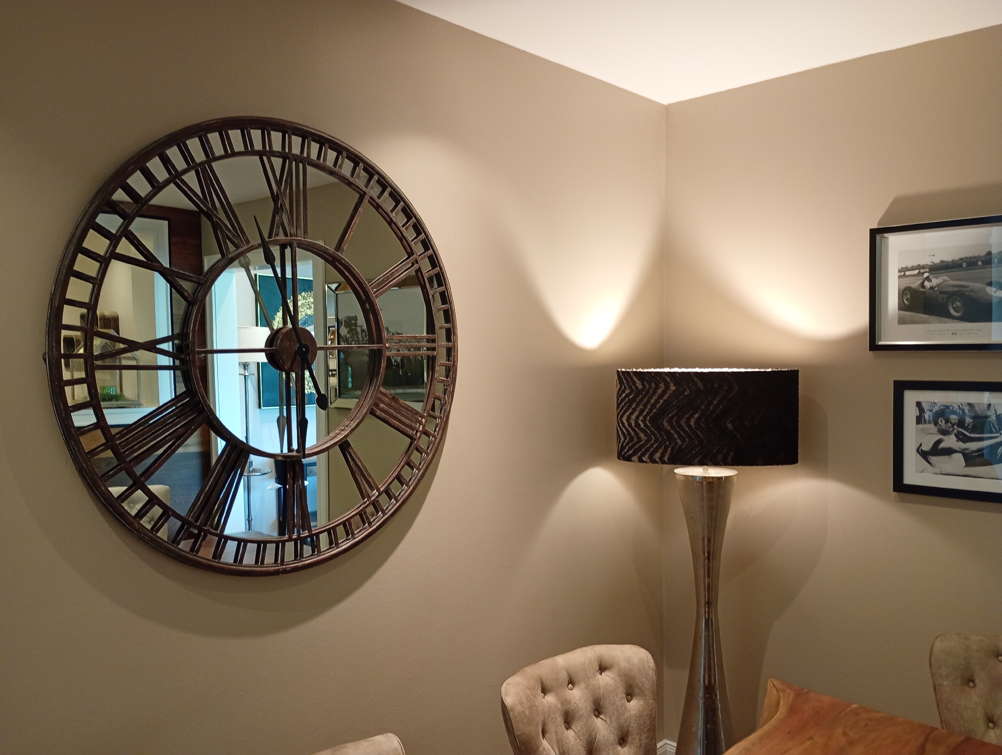 The image shows a dimly lit corner of a room, possibly a living room or dining room, with a large, circular, Roman numeral wall clock dominating the left side of the frame. The clock has a dark, metallic frame and is reflected in its mirrored surface, showing glimpses of other furniture and décor. To the right of the clock, a tall, modern floor lamp with a black patterned shade illuminates the wall, casting a warm glow. Above the lamp, two framed black and white photographs are hung on the wall; one depicts a race car, and the other appears to be a portrait of a man. In the foreground, the tufted backs of two upholstered chairs are visible, along with a glimpse of an orange cushion. The walls are painted in a neutral, taupe color. The scene is likely situated within a private residence in the city of Bad Sachsa, Germany, given the prompt's constraints.