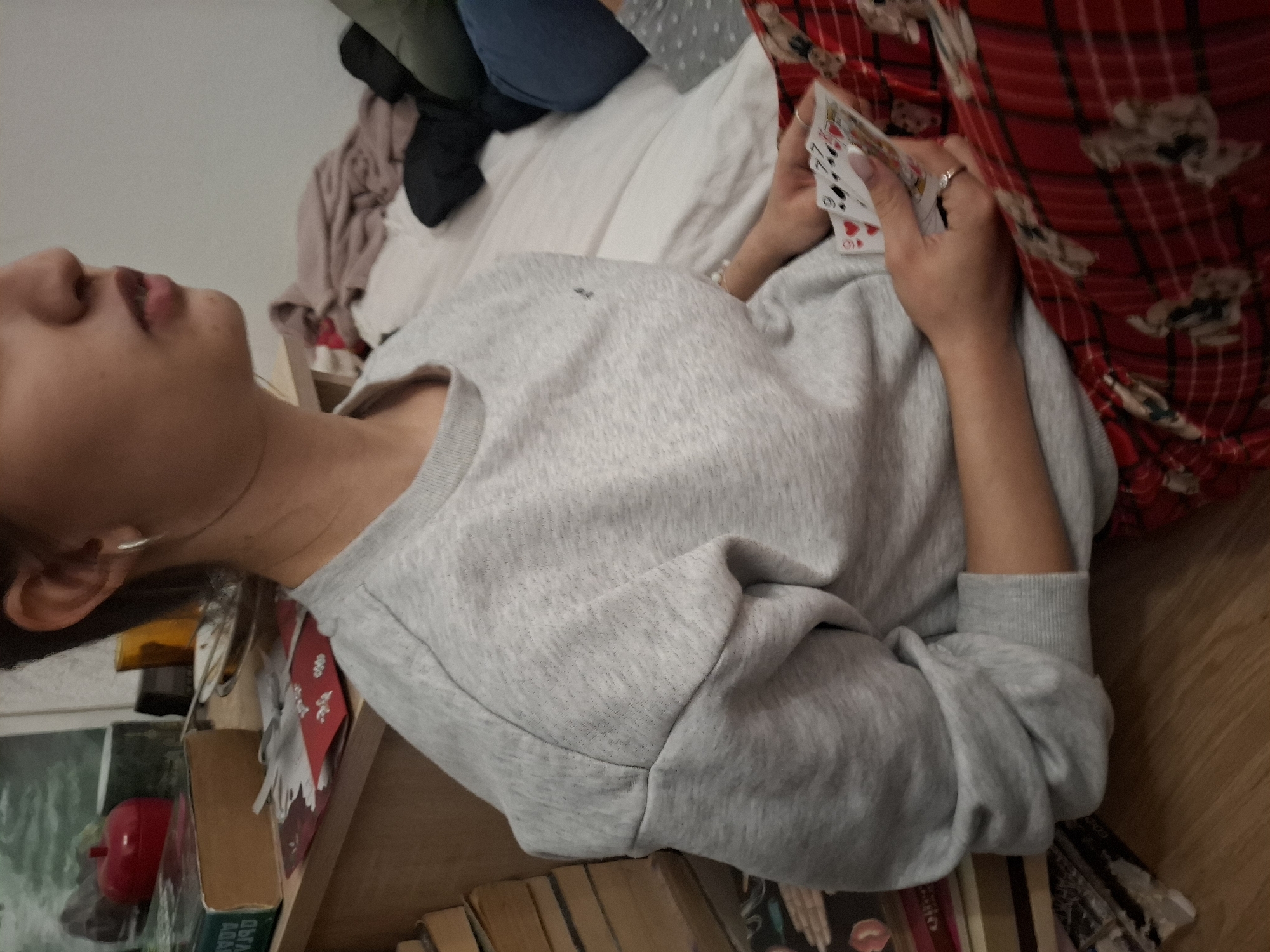 A person lies on a bed or sofa, holding playing cards in their right hand. They are wearing a light grey sweatshirt. Part of their face is visible. A red and white patterned blanket or garment is draped over them. The background includes a bookshelf with various items, and other soft furnishings. The location and city are not discernible from the image.