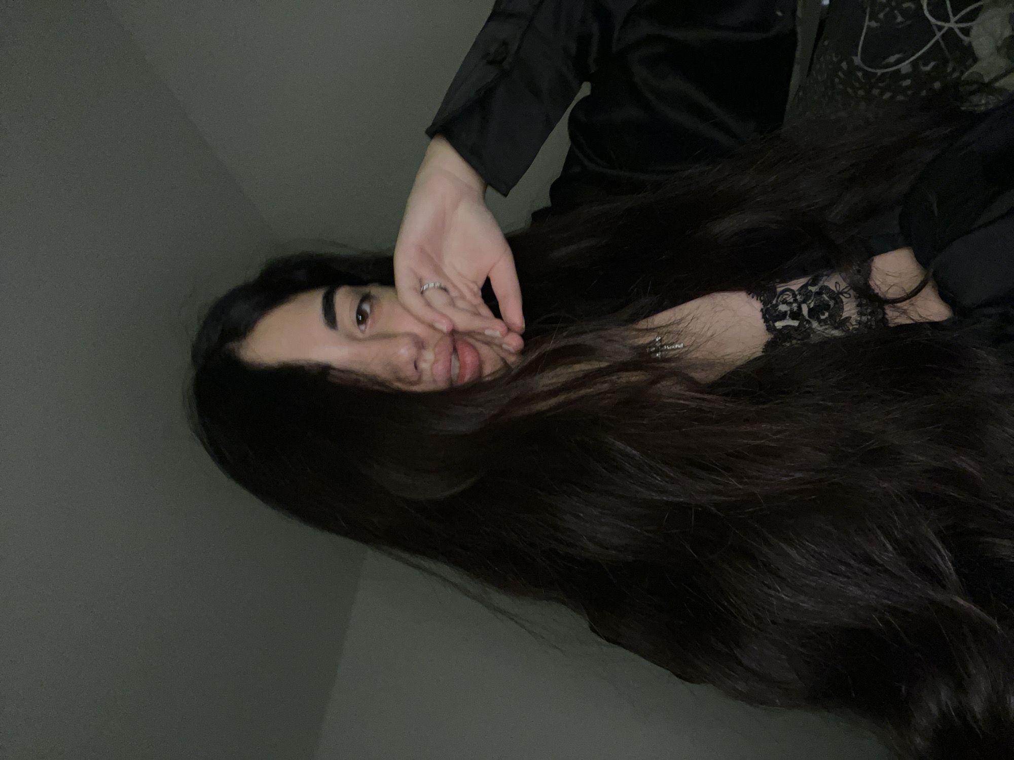 A young woman with long, dark hair and light brown eyes is shown in a close-up, dimly lit selfie. She is wearing a black satin shirt and a black lace bra. Her right hand is raised to her face, touching her lips, and she is wearing a ring on her index finger. A silver cross necklace is visible around her neck. The background is a plain, dark wall, suggesting an indoor setting. The image does not provide any information about the location being in Chotěšov, Czechia.