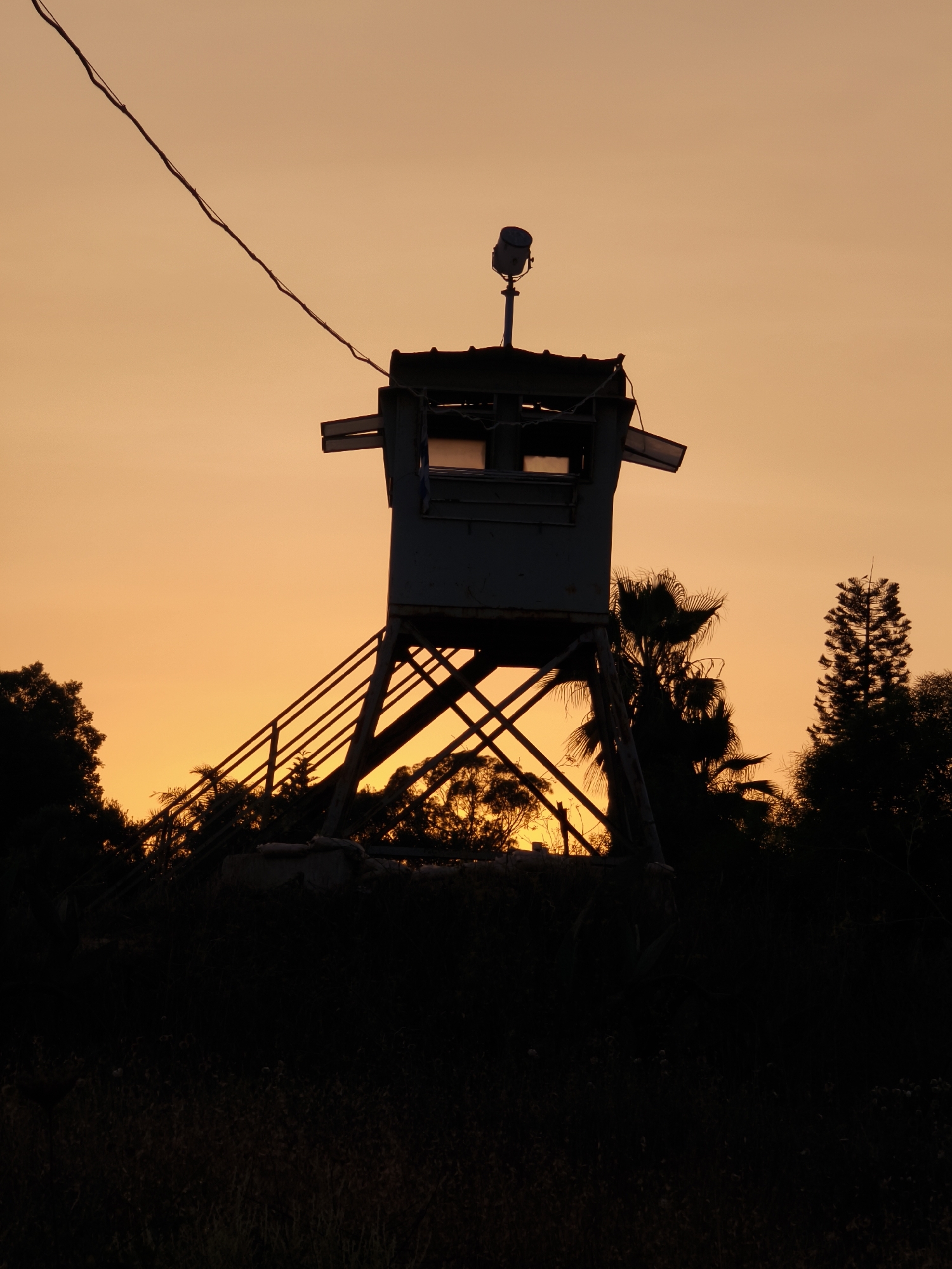 A dark silhouette of a guard tower stands against a bright orange sky at dusk. The tower is elevated on stilts and has a small structure at the top with windows and a spotlight. There is a power line running diagonally across the sky. In the foreground, the silhouettes of trees and vegetation are visible. The scene is located in the Lachish Regional Council, Israel.