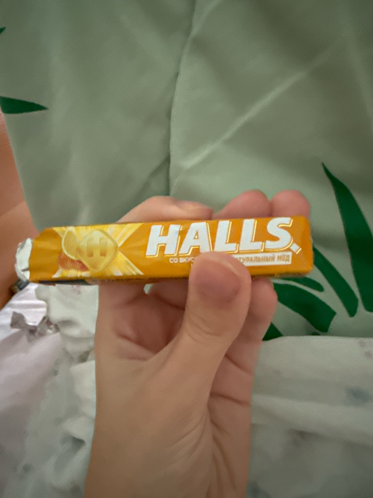 A close-up, eye-level shot shows a hand holding a Halls lozenge wrapper. The wrapper is orange with white lettering that reads "HALLS" and a smaller line of text in Russian. The lozenge itself appears to be light yellow and possibly round with a "H" imprinted on it. The background consists of a light green fabric with dark green leaf-like patterns and a white crumpled material. There is no identifiable location in the city of Zavolzhye, Russia.