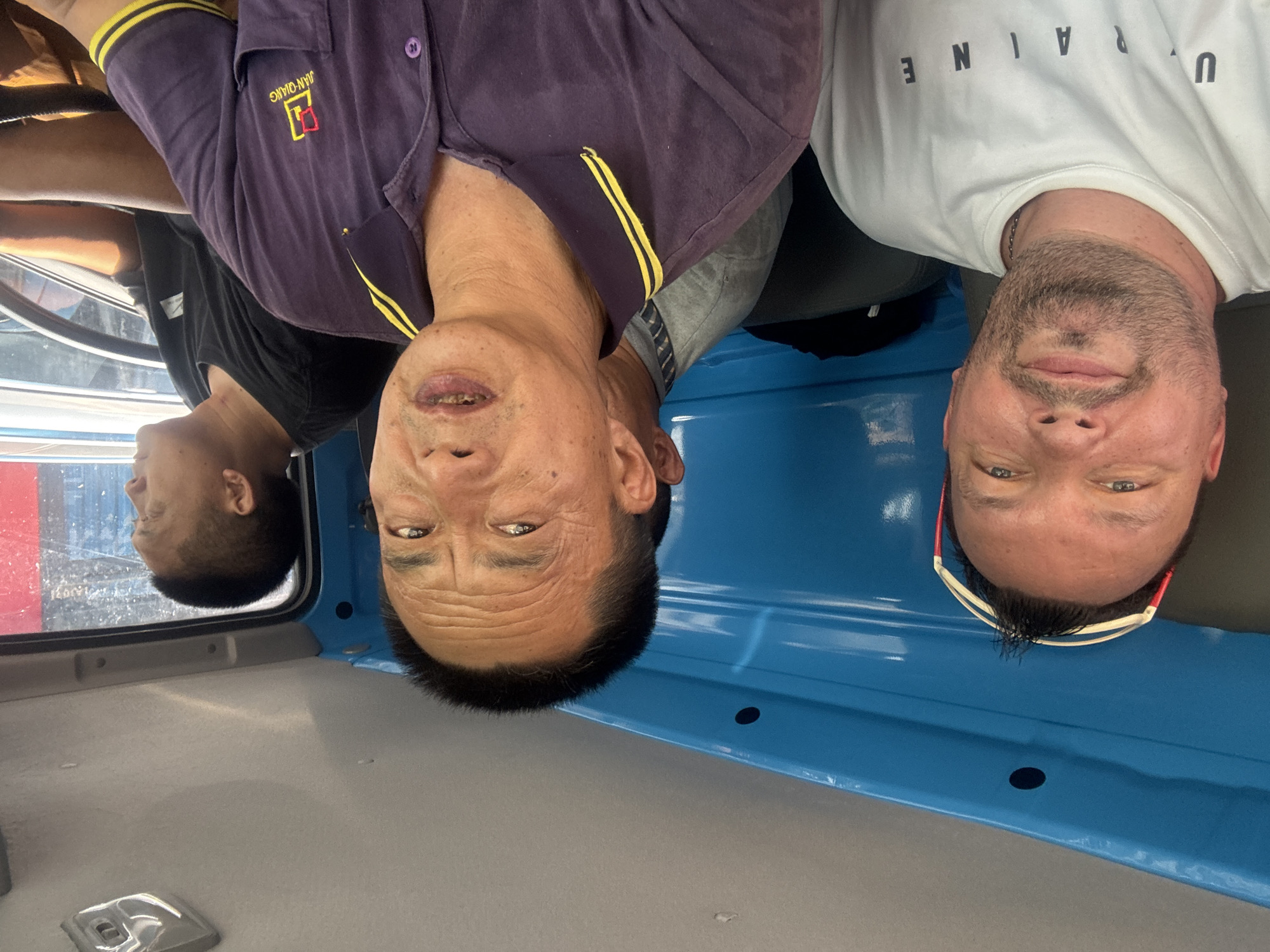 In the city of Baiyun District, China, three individuals are captured inside the cab of a vehicle. The person in the foreground, a man with short dark hair and a goatee, wears a white t-shirt with the word "UKRAINE" printed in black letters. He is looking directly at the camera, with sunglasses resting on his forehead. To his right, another man, also with short dark hair, is wearing a purple polo shirt with yellow trim and a logo that reads "JIAN-QIANG". His mouth is slightly open, suggesting he may be speaking. Behind these two men, partially visible, is a third man with short dark hair wearing a grey shirt with a striped collar. He is looking towards the left of the frame. The interior of the vehicle features a bright blue panel on the left side and a grey ceiling with a light fixture. The overall impression is a casual, candid moment captured during a journey.