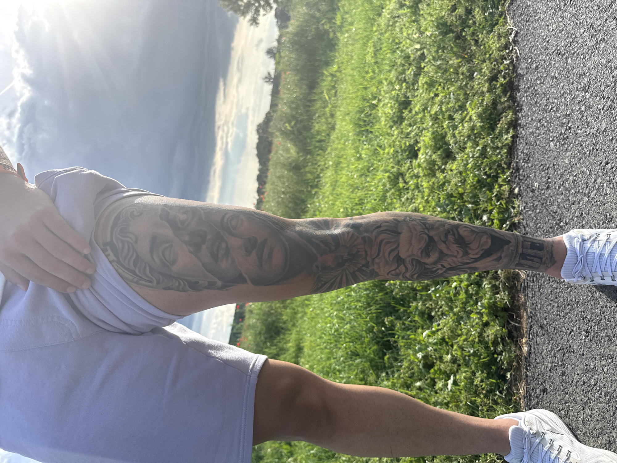 The image shows a person standing outdoors with their right arm and lower legs visible. The person is wearing light-colored shorts, a light-colored shirt, and white sneakers. The right arm is extensively tattooed with a design that includes a human face and other ornate patterns. The person is standing on an asphalt path, with grass visible on one side of the path. The background shows trees, a cloudy sky, and a bright sun. The location is identified as Elhovo, Bulgaria.