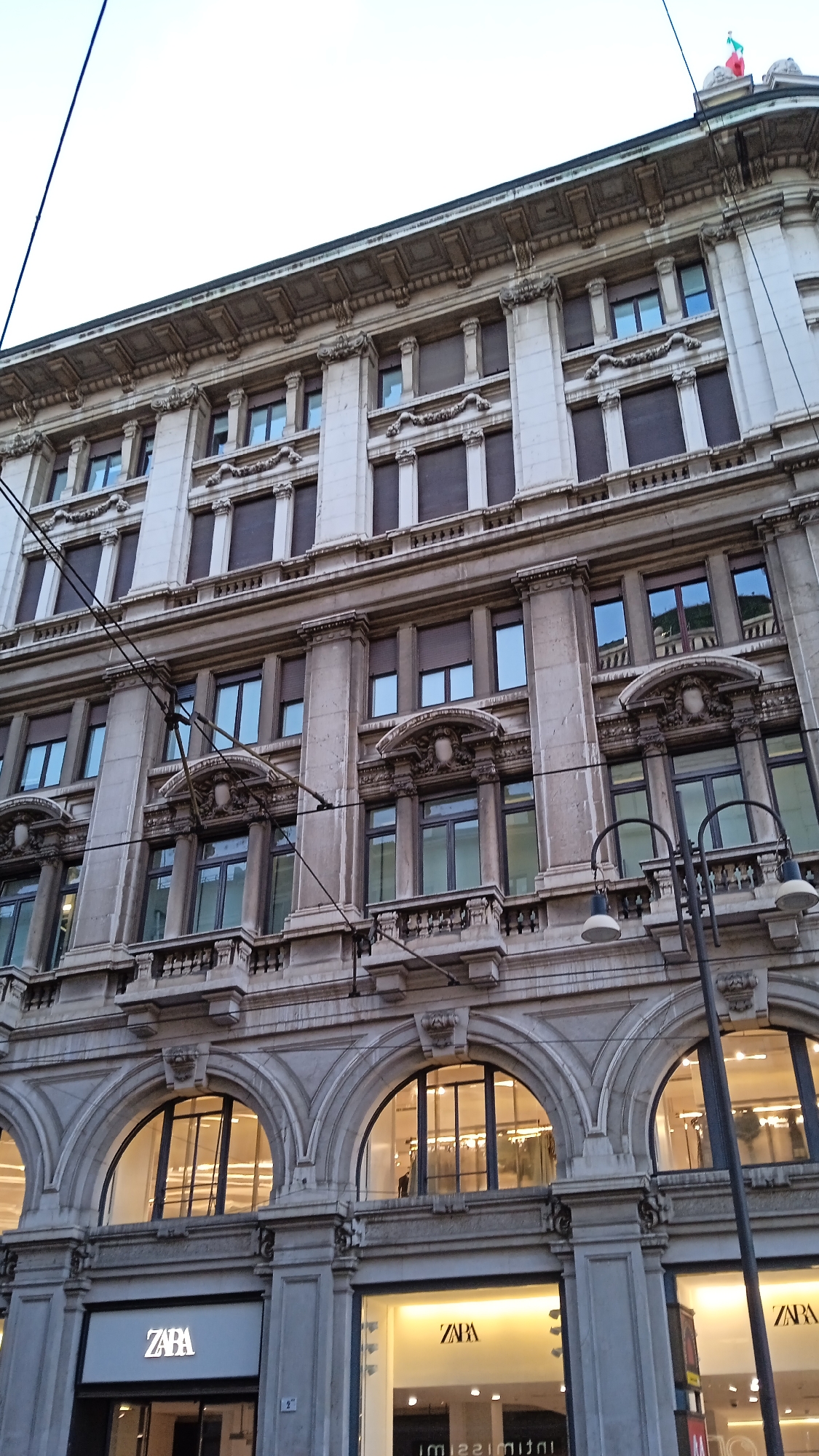 This is a photograph of the Zara store located at Corso Vittorio Emanuele II in Milan, Italy. The image captures the upper levels of a multi-story building with ornate architectural details, including columns, cornices, and decorative carvings. Several windows with dark shutters are visible on the upper floors. The lower level of the building features large arched windows, behind which the brightly lit interior of the Zara store can be seen. The Zara logo is prominently displayed on the facade of the store in two locations. The street level features storefronts and what appear to be display windows. The sky above is overcast. There are no visible people in the photograph.