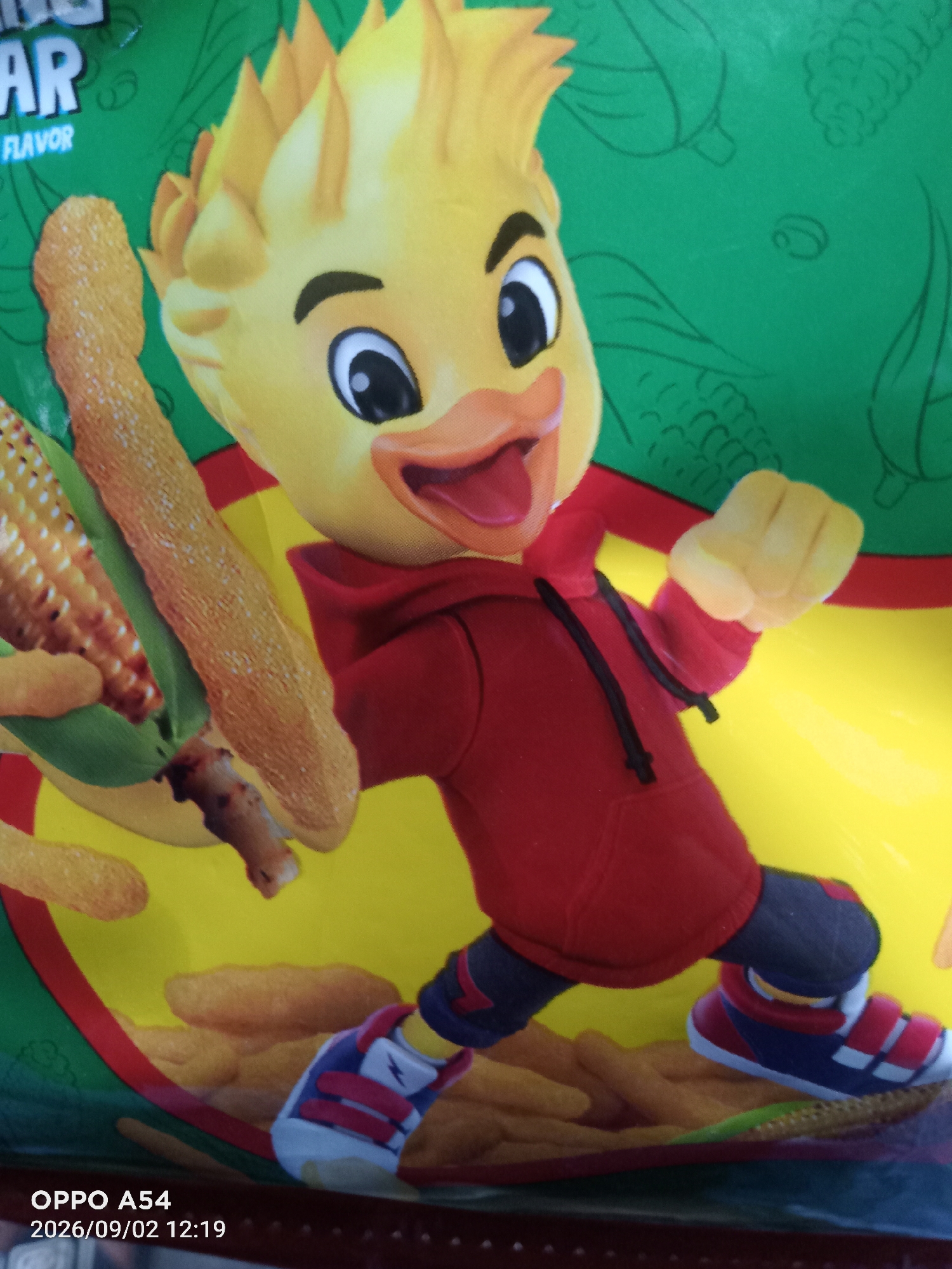 The image is a close-up, eye-level shot of a snack bag featuring a cartoon duck character holding a piece of corn and a crunchy snack. The duck has a yellow head with spiky hair, large black eyes, an orange beak, and is wearing a red hoodie and dark pants. It is depicted in a dynamic pose, with one fist raised and the other arm extended, holding the corn cob and snack. The background is predominantly green with line drawings of corn cobs and other produce, and a bright yellow area towards the bottom. In the lower left corner, a timestamp reads "OPPO A54 2026/09/02 12:19". The image was taken in the city of Jambi, Indonesia.