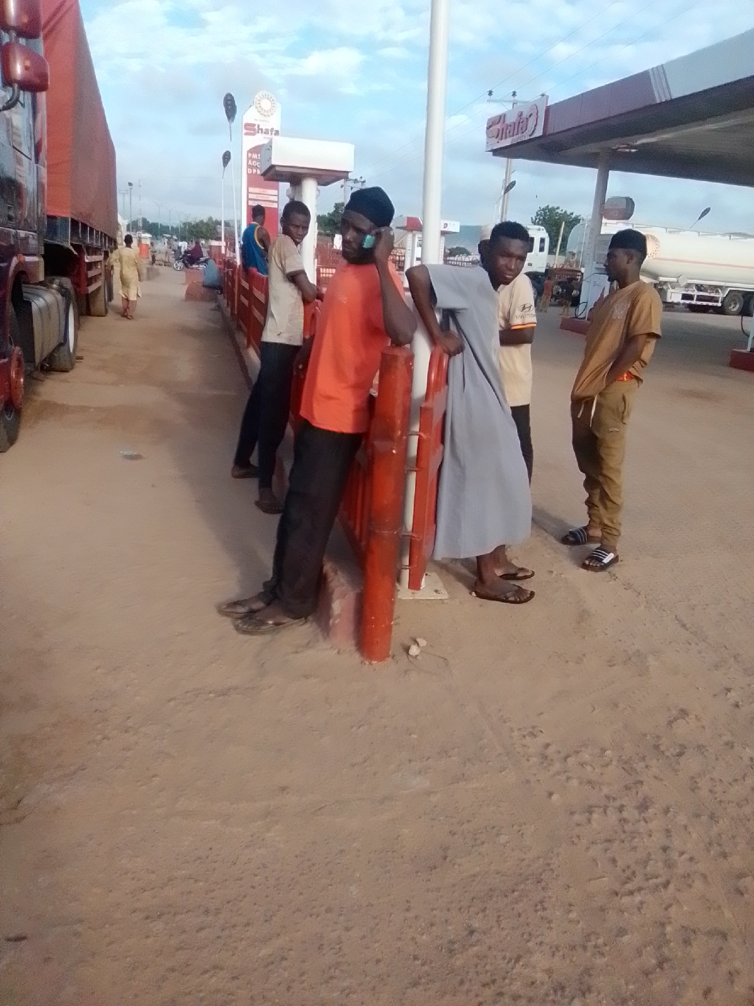 In Kwadon, Nigeria, this image captures a group of young men gathered at a gas station. A large red truck dominates the left side of the frame, its trailer covered with a tarp. Three young men stand near a red barrier in the foreground. One, wearing an orange shirt and black pants, is talking on a mobile phone. Another, in a grey robe, leans on the barrier. The third, in a tan shirt and brown pants, stands further back. Behind them, a gas station sign with "Shifa" written on it is visible. The ground is sandy and uneven, with tire tracks suggesting vehicle movement. The sky is partly cloudy, indicating it is daytime.