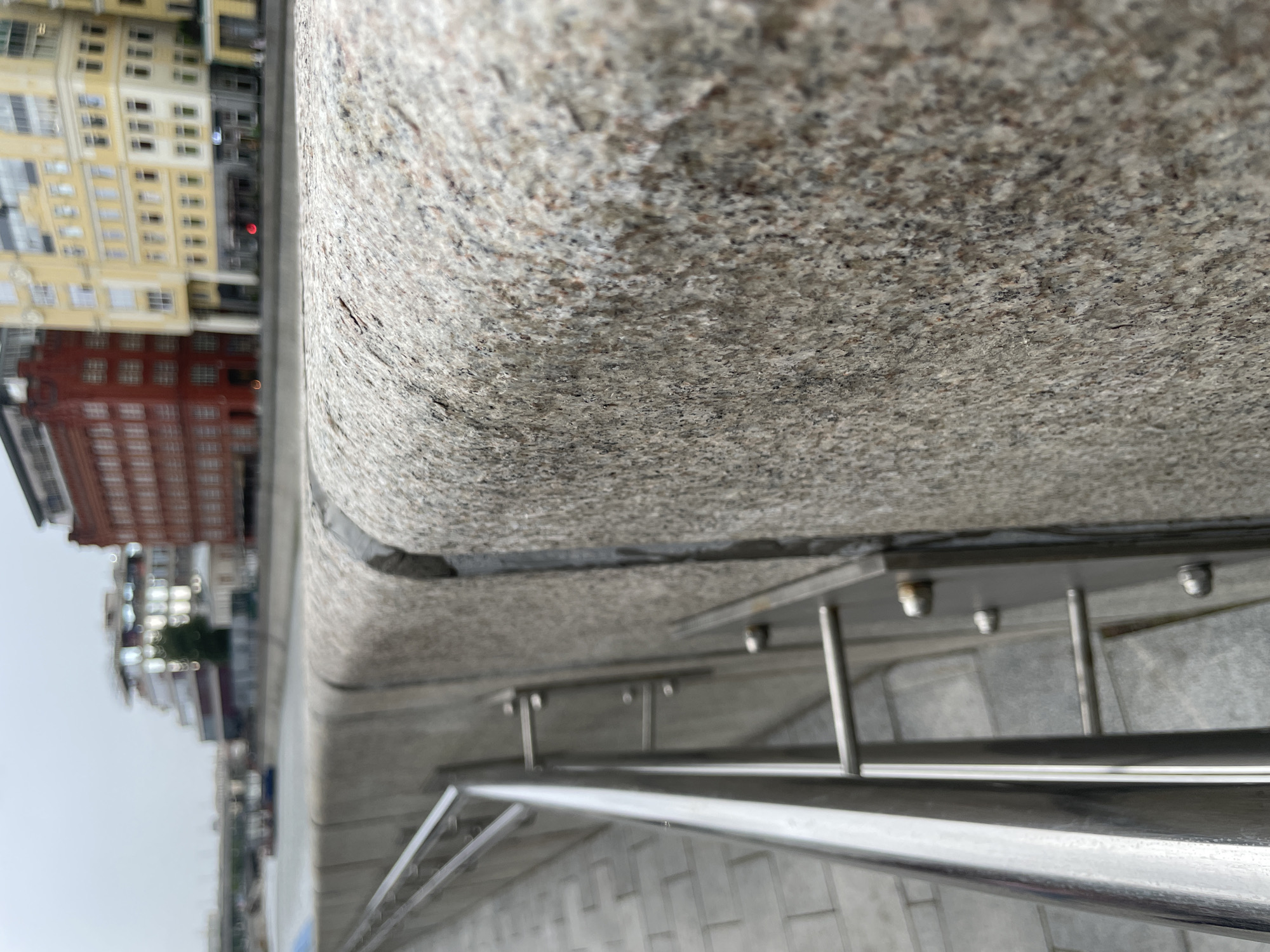 This is a close-up, eye-level, exterior, medium shot photograph taken on an overcast day. The focal point is a coarse-grained, light gray granite wall that extends from the bottom right corner of the frame to the left side. The wall has a rounded top edge, with a darker seam running along its length. To the left of the granite wall is a metal railing that runs diagonally up and to the left, appearing to follow a descending path. The railing is made of polished metal poles and vertical supports. The ground beneath the railing is paved with gray square tiles. In the background, a river or canal is visible, with a boat on the water. Beyond the water, a cityscape is present, featuring buildings with various architectural styles and colors, including a red brick building and a yellow building. The sky is overcast and light gray.