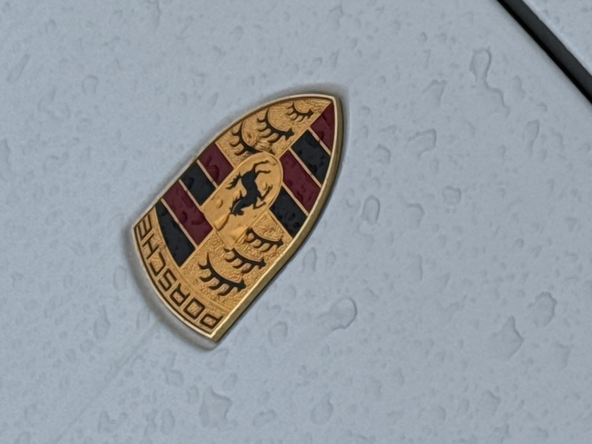The image shows a close-up of a Porsche emblem affixed to a car. The emblem features a rearing horse at the center, surrounded by stylized antlers above and the word "PORSCHE" below. The background of the emblem is divided into quarters with alternating black and red stripes. The car's surface around the emblem appears to be light gray and is covered with water droplets, suggesting it has recently rained. The image does not provide enough information to determine the location in Plovdiv, Bulgaria or any interactions.