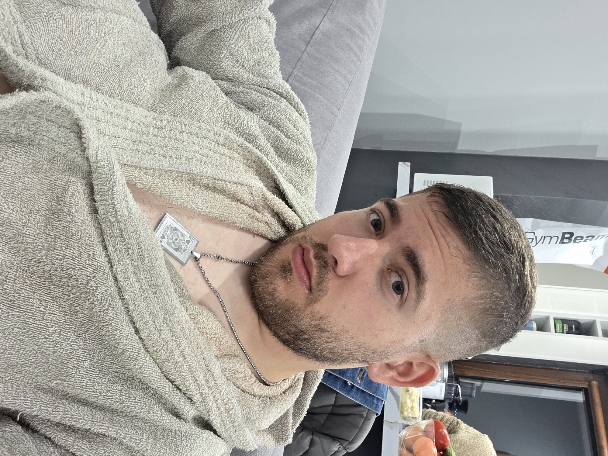 A man with a short, styled haircut and a beard is wearing a light-colored bathrobe and a silver necklace with a square pendant. He is reclining on a gray couch or sofa. In the background, there is a white cabinet or shelf unit with various items on display, including a GymBeam-branded product. The setting appears to be indoors, possibly a residential space. There's not enough information to determine if this is taken in Yagodovo, Bulgaria.