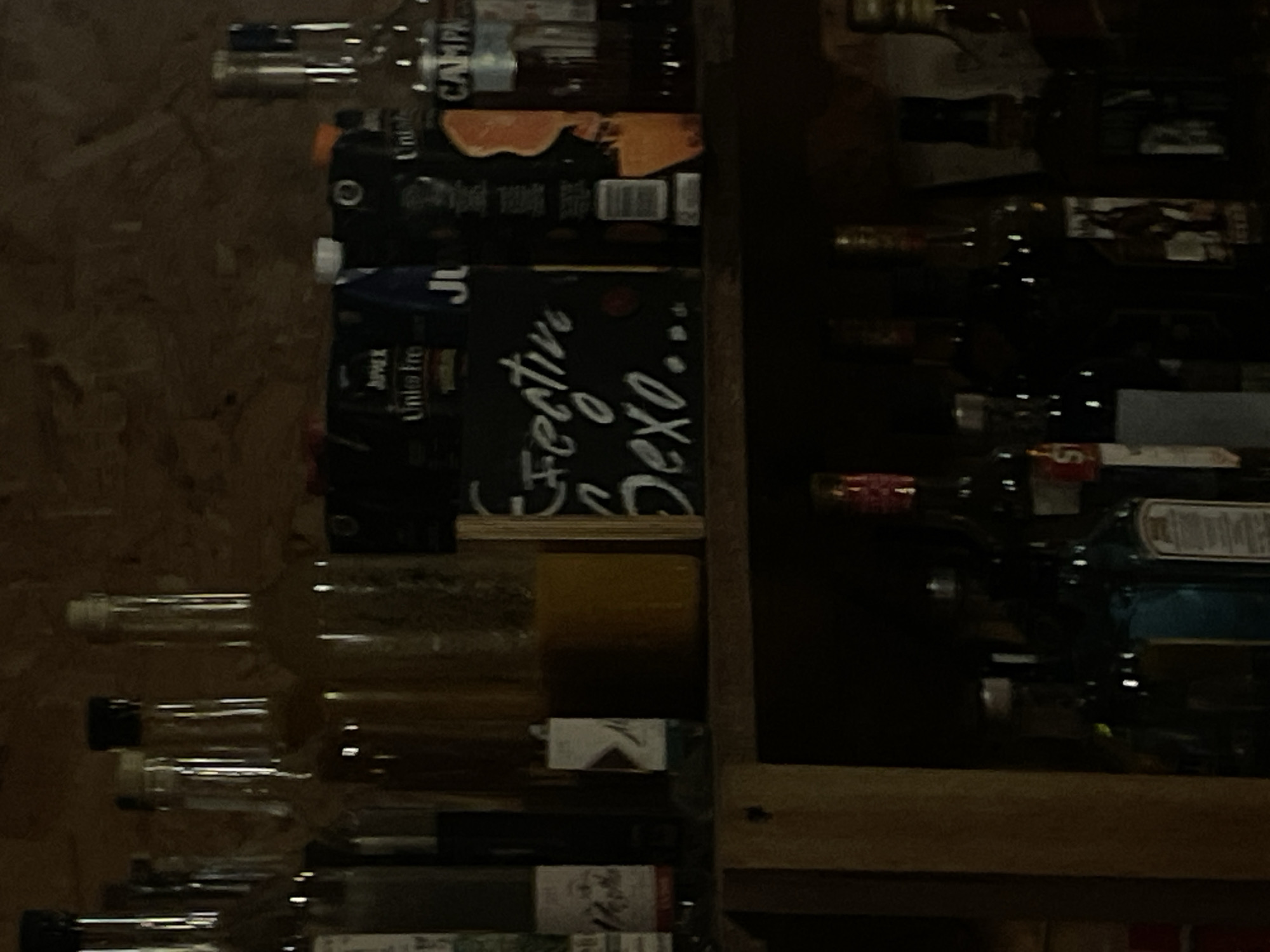 The image shows a shelf stocked with various bottles and containers, likely alcohol or mixers. Labels like "Campari", "Unifre", and "Festive Sexo..." are partially visible. The lighting is dim, and the image is somewhat blurry. Based on the labels and the context, it's likely a bar or restaurant in Puerto Escondido, Mexico. There is no presence of people in this image.