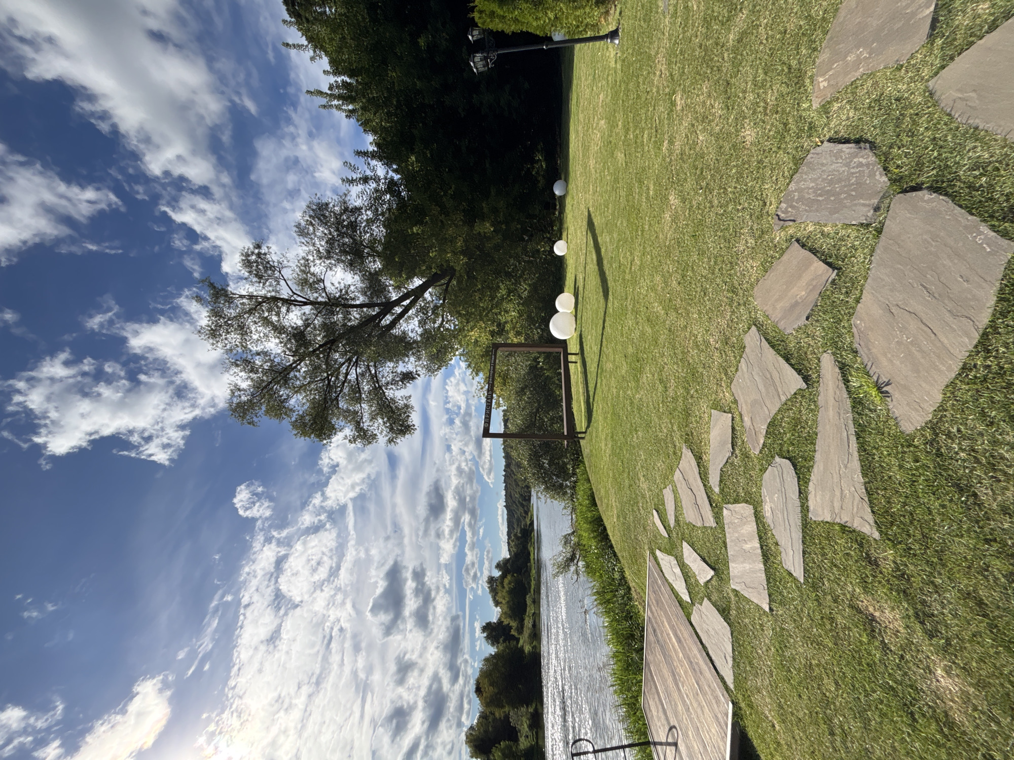 This is a photograph of a waterside lawn in Zvenigorod, Russia, under a partly cloudy sky. In the foreground, a wooden deck leads to a path of large, irregular flagstones laid on a green, mown lawn. Further into the lawn, a large, dark wooden frame stands with a bench in front of it. To the right of the frame, three white spherical decorative lights are placed on the grass. A black lamp post stands to the right of the lights, and a dense line of green trees forms the backdrop of the lawn. To the left of the lawn, a wide, calm river flows, with green foliage and trees lining its far bank. The sun is visible in the upper left corner of the sky, casting a slight lens flare. There are no people or animals visible in the image.