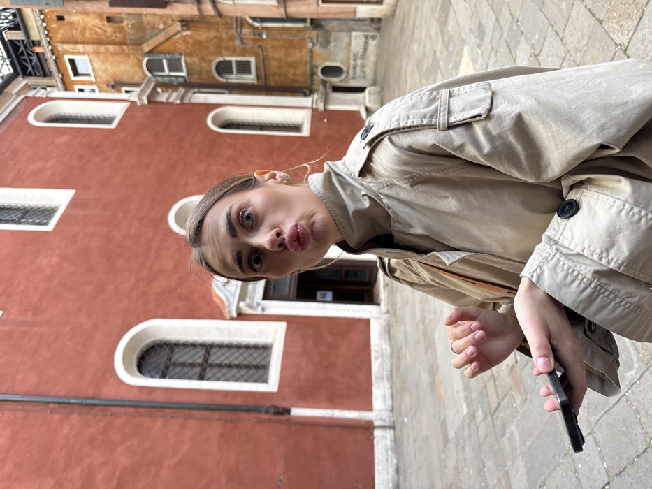 A single female subject is depicted outdoors, captured from the mid-torso up. She has light brown hair, pulled back, and is wearing a light beige trench coat featuring shoulder epaulets, a left sleeve pocket, and dark cuff buttons. Her facial expression shows wide eyes and puckered lips. She holds a dark-colored smartphone horizontally in her left hand.
The background features a prominent, terracotta-red building with multiple arched windows framed in white, and a white-trimmed ground-level entrance. A black downspout runs vertically on its left side. To the right, a narrower, taller building with an orange-brown stucco facade and arched windows is visible, featuring metal structural elements at its apex. Graffiti, including the name "LUIGI MANZONI," is present on the lower section of the orange-brown building's wall. The ground is paved with stone. This scene is located in Campo San Giacomo dall'Orio, Venice, Italy. The lighting suggests an overcast day.