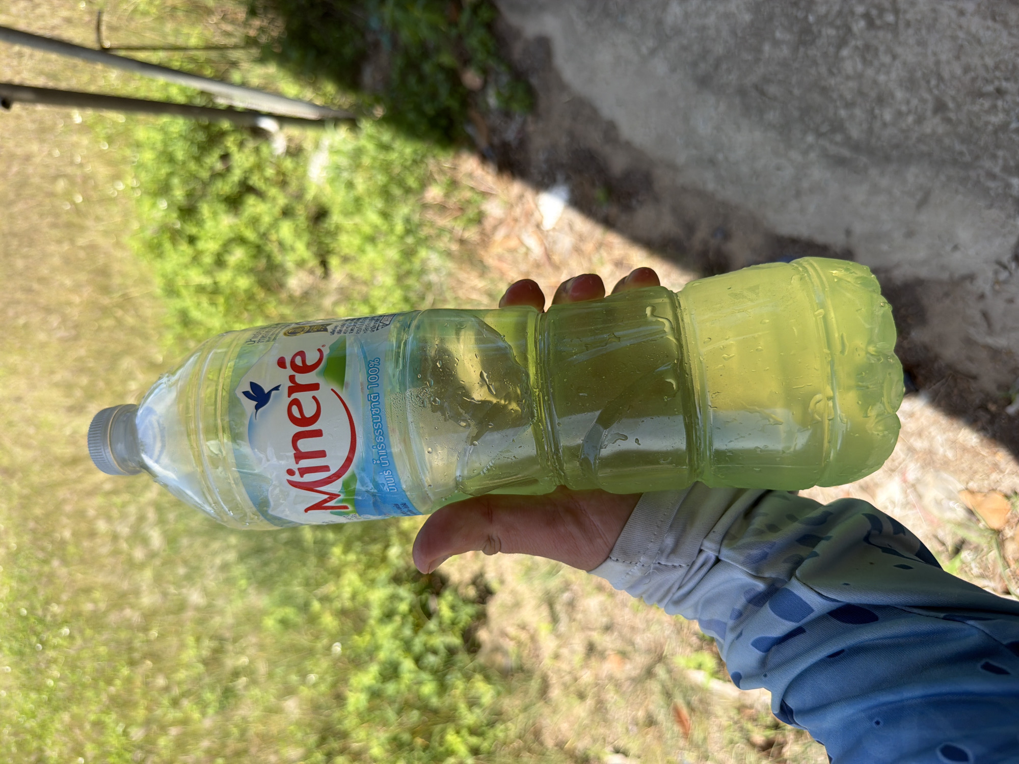 A person's hand, wearing a blue and white patterned long-sleeved shirt, holds a clear plastic bottle of Mineré mineral water. The bottle is about two-thirds full of a yellow liquid and has condensation on its exterior. The bottle's label features a red "Mineré" logo with a blue bird silhouette and Thai text below. The background consists of blurred green foliage and a concrete surface, suggesting an outdoor setting. The lighting indicates it is daytime. The location is Baan Bo Phut, Thailand.