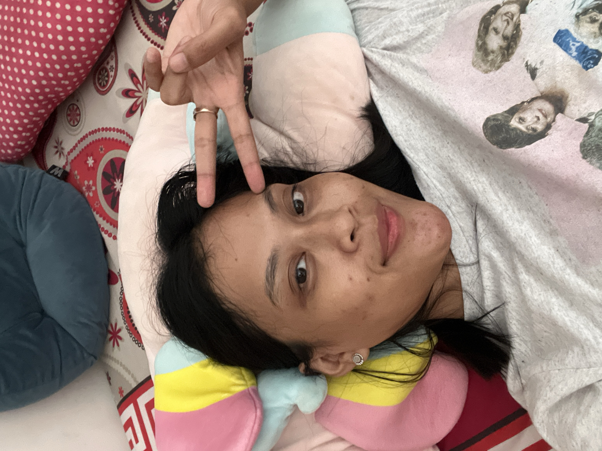 In Kecamatan Pati, Indonesia, a young woman with dark hair and a light complexion is lying on a bed. She is looking directly at the camera, her right hand raised with two fingers extended in a peace sign. She is wearing a light-colored t-shirt with a graphic of four women. She is lying on a bed with colorful pillows and a patterned duvet. The scene appears to be indoors, possibly in a bedroom.