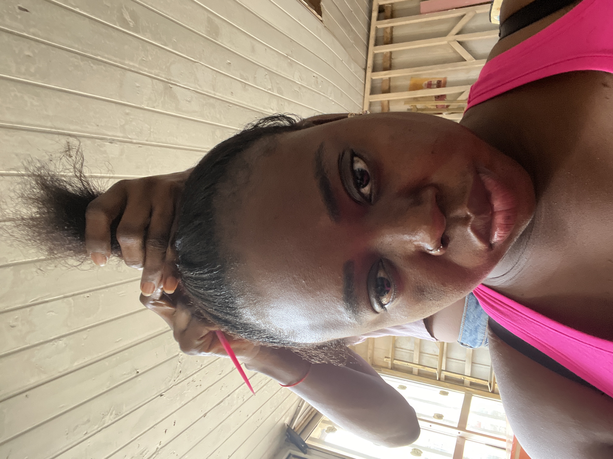 A close-up, overhead shot shows a young Black woman from the chest up as her hair is being braided. She is wearing a bright pink tank top and has a nose ring. Her hair is pulled back and is in the process of being styled. A pink comb is visible in her hair, held by a person whose hands and arm are in the background. The background consists of a wooden ceiling and a partial view of a room with wooden beams and windows. The lighting is somewhat dim.