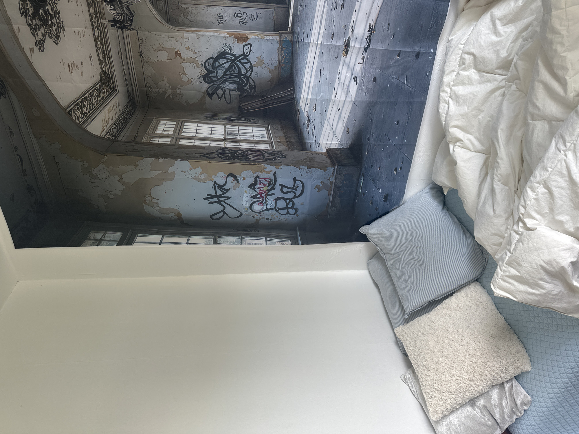 The image depicts an interior scene featuring a bed with several pillows and a white duvet in the foreground. Behind the bed, a large mural covers the wall, creating the illusion of an abandoned, ornate room with peeling wallpaper, graffiti, and debris on the floor. Sunlight streams in through large windows, casting shadows. The mural's style suggests an urban decay aesthetic. The setting is not identified as being in Białystok, Poland.
