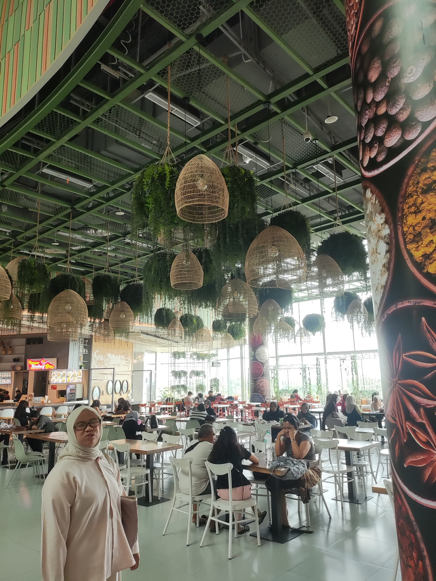 The image depicts the interior of a food court in Lambangjaya, Indonesia. The space is characterized by a high ceiling with exposed green metal beams and a mesh grid. Hanging from the ceiling are numerous woven basket-like lamps and a dense collection of artificial plants, creating a suspended garden effect. The food court features multiple tables and chairs arranged in dining areas. In the foreground, a woman wearing a light pink hijab and matching outfit is the primary subject, looking towards the camera. In the background, other patrons are seated at tables, engaged in eating or conversation. A large, decorative pillar with images of spices is visible on the right side of the frame. The overall ambiance suggests a bustling, modern dining establishment.