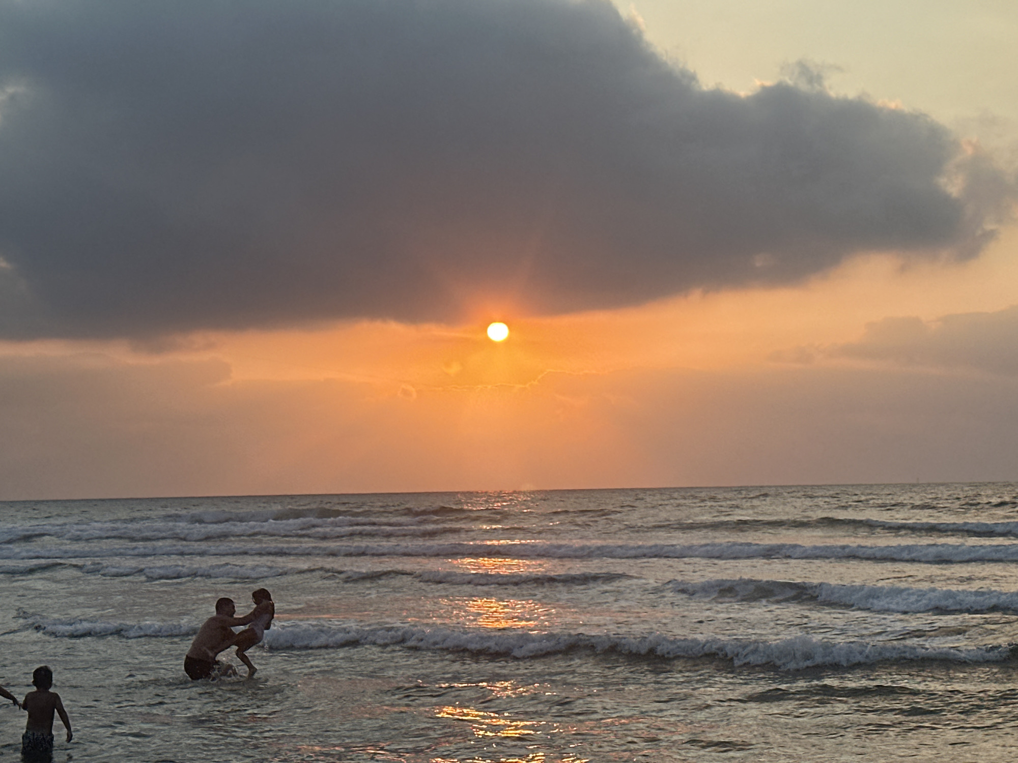 At sunset, on the coast of Haifa, Israel, a group of people are enjoying the ocean. The sun, a bright orange orb, is partially obscured by dark clouds, casting a warm glow over the water. Waves gently lap at the shore, and two children and a man are playing in the surf. The man is holding a young girl, lifting her in the air. Another child stands in the shallow water nearby. The scene is tranquil, with the soft light and the sound of the waves creating a peaceful atmosphere.