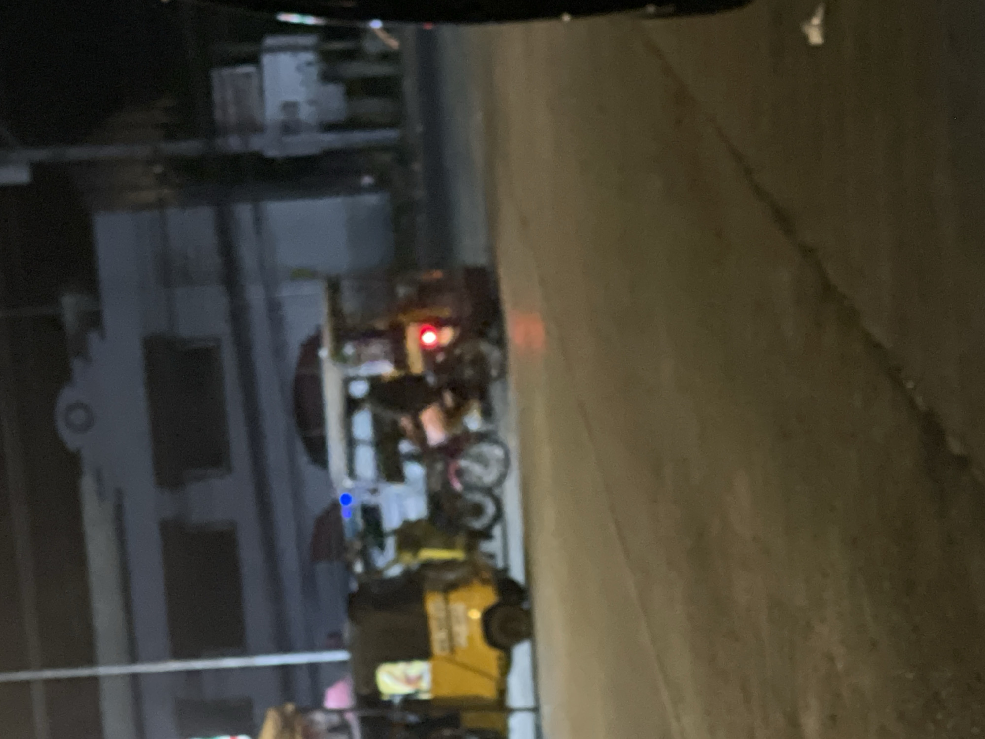 An out-of-focus, night-time street scene in Davao City, Philippines, captures a yellow auto-rickshaw, also known as a " motorela" in the Philippines, on the left side of the frame. The motorela has a black canopy and appears to be carrying passengers or goods, indicated by a yellow and red graphic on its side. In the background, to the right of the motorela, a red motorcycle is partially visible, with a rider on it. A white, bus-like vehicle is situated further back, behind the motorcycle. The street is paved and appears wet, suggesting recent rain. The overall ambiance is dimly lit, with artificial lighting from streetlights and vehicles contributing to the low visibility. The buildings in the background are indistinct, with a few windows visible. The image's blurriness suggests it was taken while in motion, possibly from another vehicle.