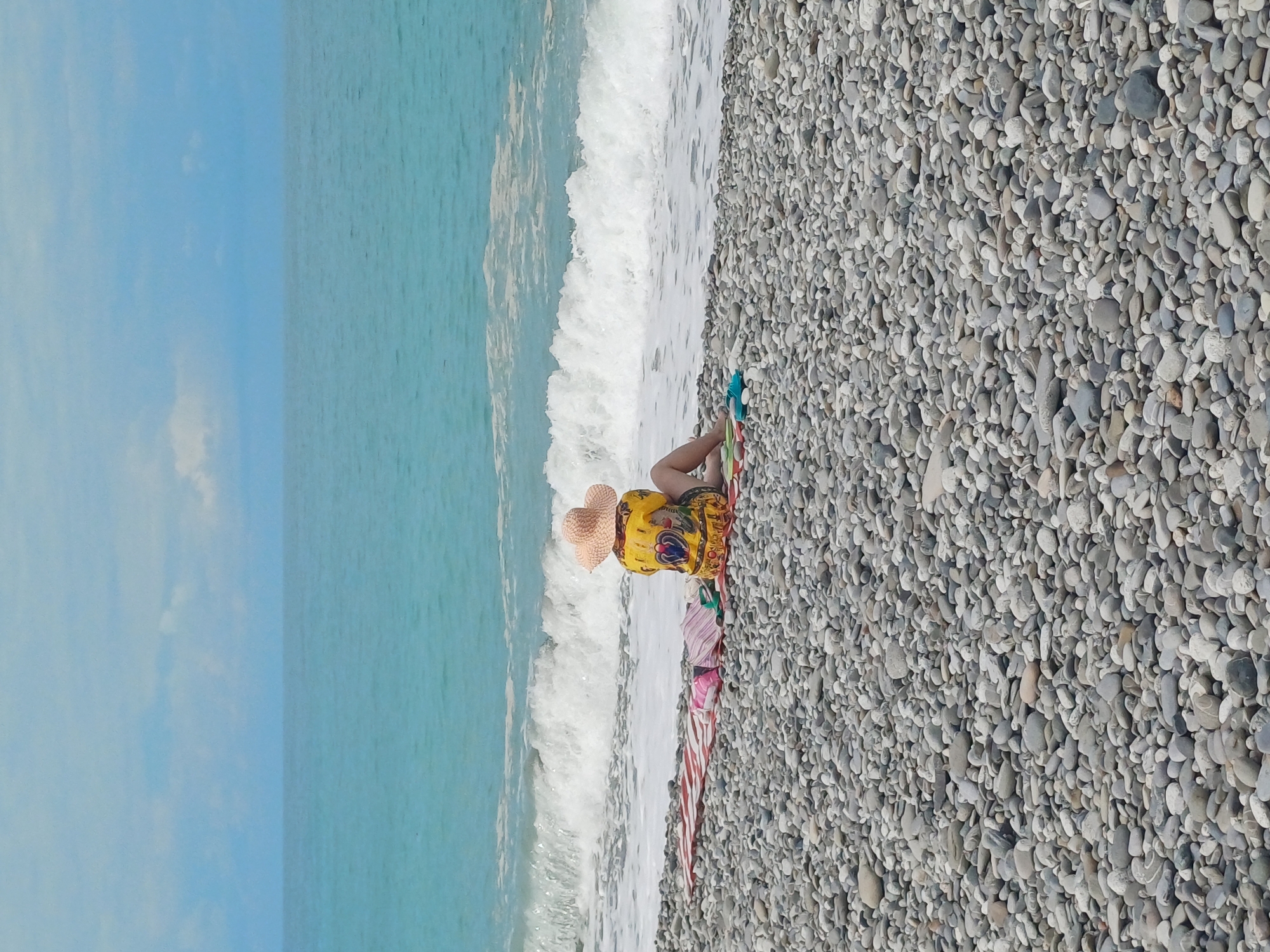 A person sits on a pebble beach in Sochi, Russia, facing the sea. The person is wearing a straw hat and a yellow shirt with a colorful pattern. They are sitting on a striped towel with other folded towels and a pair of blue flip-flops next to them. The sea is turquoise blue with white waves breaking on the shore. The sky is a pale blue with scattered clouds. The pebble beach extends out of frame.