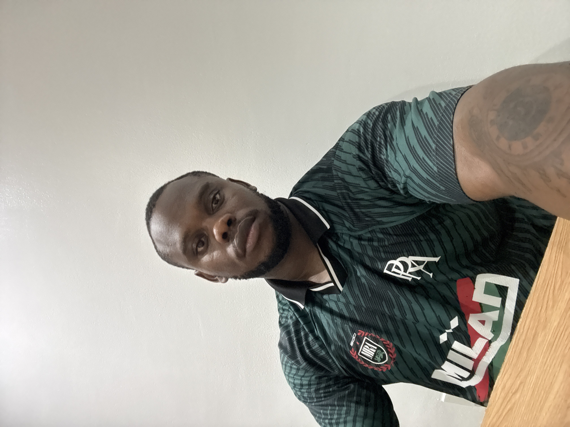 The image presents a portrait of a Black man, captured indoors from a slightly low angle. He has short dark hair and a well-trimmed beard, looking directly at the camera with a neutral expression. The photograph is rotated counter-clockwise, positioning his head on the left side of the frame and his body extending towards the lower right.
He is wearing a dark green polo shirt featuring a subtle, striped pattern. The shirt has a black collar accented with white stripes. Prominent branding is visible on his attire: a circular crest with red and green elements and what appears to be "MBE" in the center is on his left chest. Above his right chest, white letters spell "RRA". Across the front of the shirt, large white lettering forms the word "MILAN," integrated with red and green accents, likely referencing the Italian flag colors.
A significant, dark, circular tattoo, possibly resembling a clock face, is visible on his left arm (appearing on the right side of the image due to the angle). A light wooden surface, suggestive of a table or desk, is partially visible along the right edge of the frame. The background is a plain, light-colored wall, indicating a simple indoor setting. The lighting is bright and even, consistent with typical indoor illumination. This casual portrait was taken in Iddo Tudunwada, Nigeria.