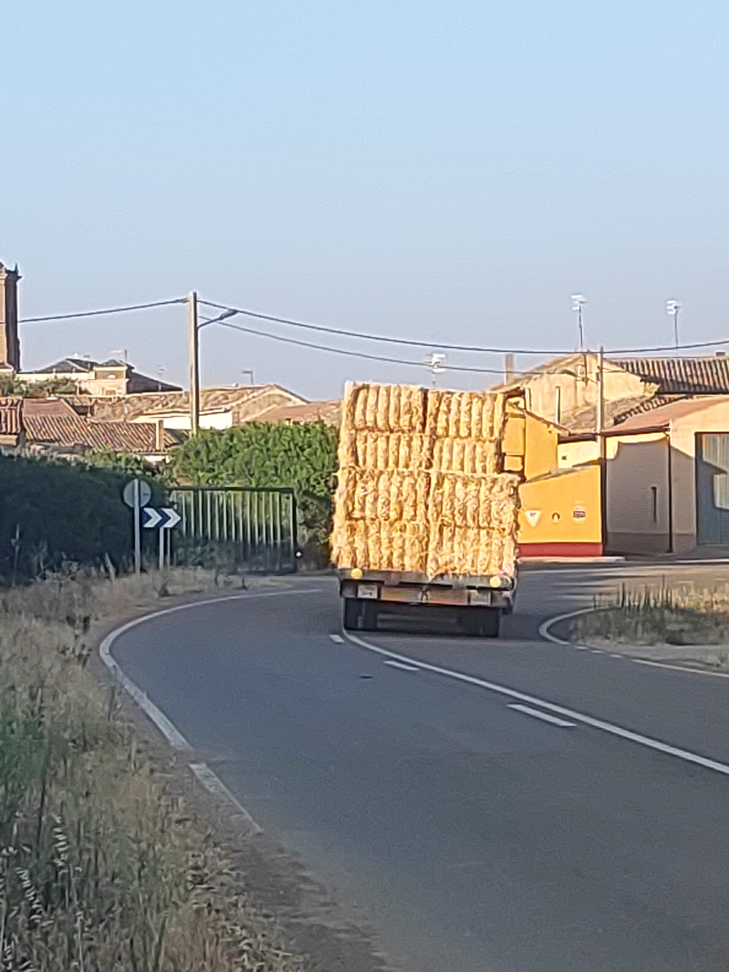 A flatbed truck carrying a large stack of hay bales turns on a road in Autillo de Campos, Spain. The hay bales are tightly packed and extend upwards, creating a towering load. The truck is dark colored, and its tires are visible. The road is asphalt and curves to the right, with white lane markings. On the left side of the road, there is a grassy verge and a blue and white directional sign with an arrow pointing to the right. Beyond the verge, there are trees and buildings with red tile roofs. The sky is clear and blue. The buildings appear to be a mix of residential and agricultural structures, with some appearing to be older or more rustic in style. One building is painted yellow and has a red base. There are power lines visible in the background, stretching across the sky. The overall scene suggests a rural setting, likely during daytime.