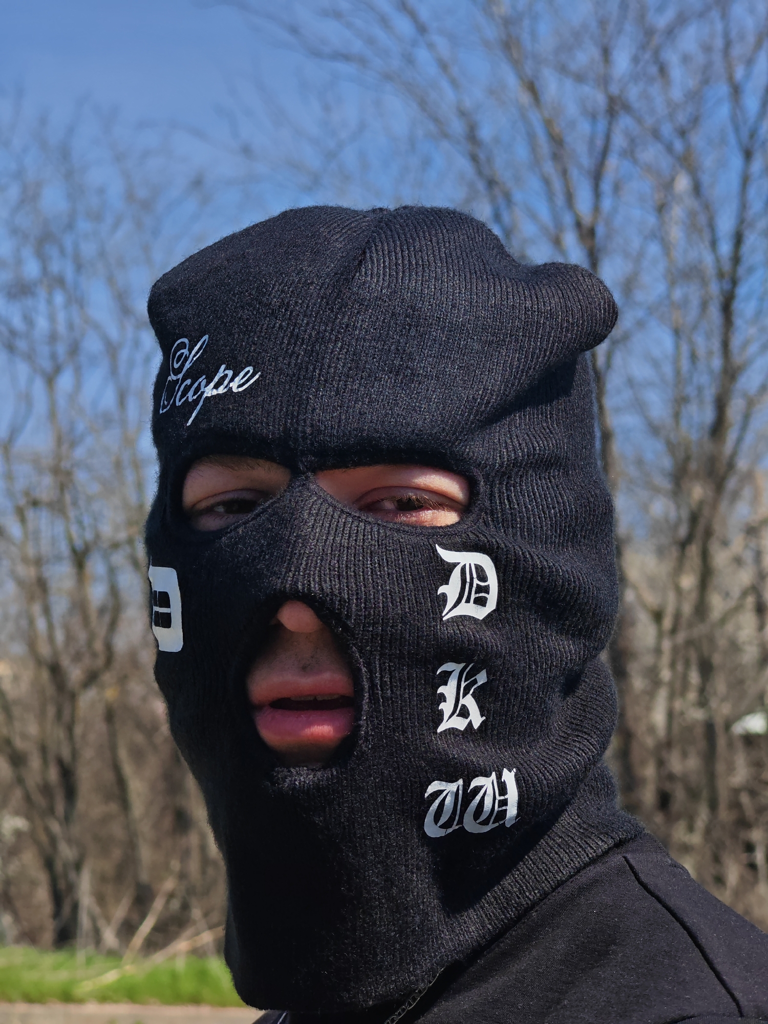 A single individual is depicted in a medium close-up, facing directly towards the observer in an outdoor setting, potentially within or on the outskirts of Shumen, Bulgaria. The subject wears a black knitted balaclava covering the entire head and face, with cutouts for the eyes and mouth. The balaclava features white embroidered text: "Scope" in script on the forehead, an incomplete white square symbol resembling "SH" on the left cheek (viewer's right), and three stacked letters "D", "K", "W" in an Old English font on the right cheek (viewer's left). The subject's dark eyes are visible through the eyeholes, and the mouth is slightly open, revealing pink lips and a glimpse of upper teeth. The individual is also wearing a black garment, possibly a hooded sweatshirt, beneath the balaclava. The background is out of focus, consisting primarily of bare deciduous trees with thin branches against a clear blue sky. A narrow strip of ground, appearing to be an unpaved surface or dry vegetation, is visible at the bottom of the frame. The scene is illuminated by natural daylight.
