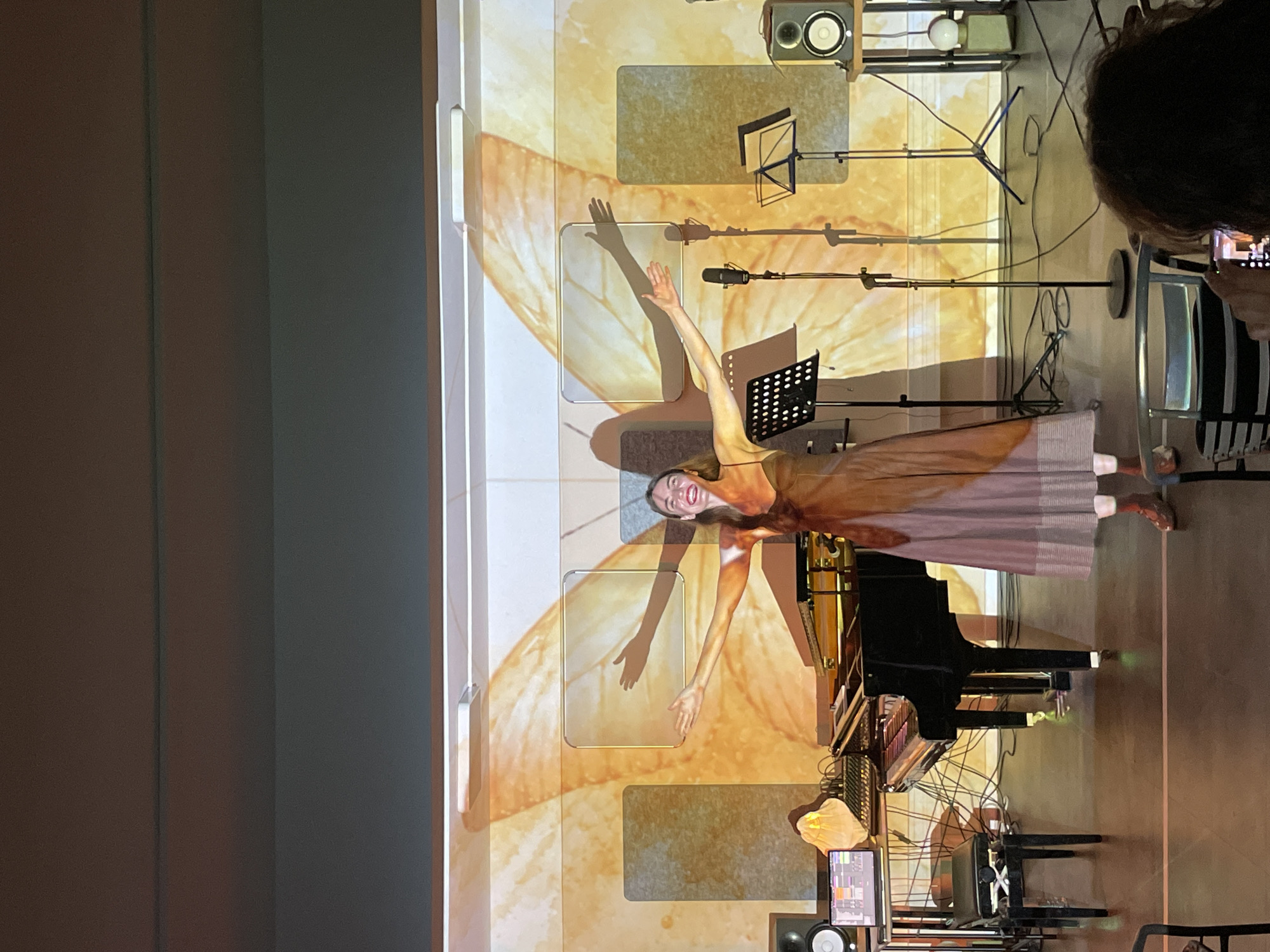 A woman stands in front of a grand piano with her arms raised against a projected backdrop featuring a large moth wing pattern and various rectangles. She is wearing a light-colored dress that blends with the backdrop's tones. The location is an interior space that looks to be used for performance and recordings. The image was likely captured during a performance. There is musical equipment visible: microphones, music stands, and a sound system. A person out of focus in the upper right corner is taking a photograph. The city of Tivat, Montenegro is not apparent in the image.