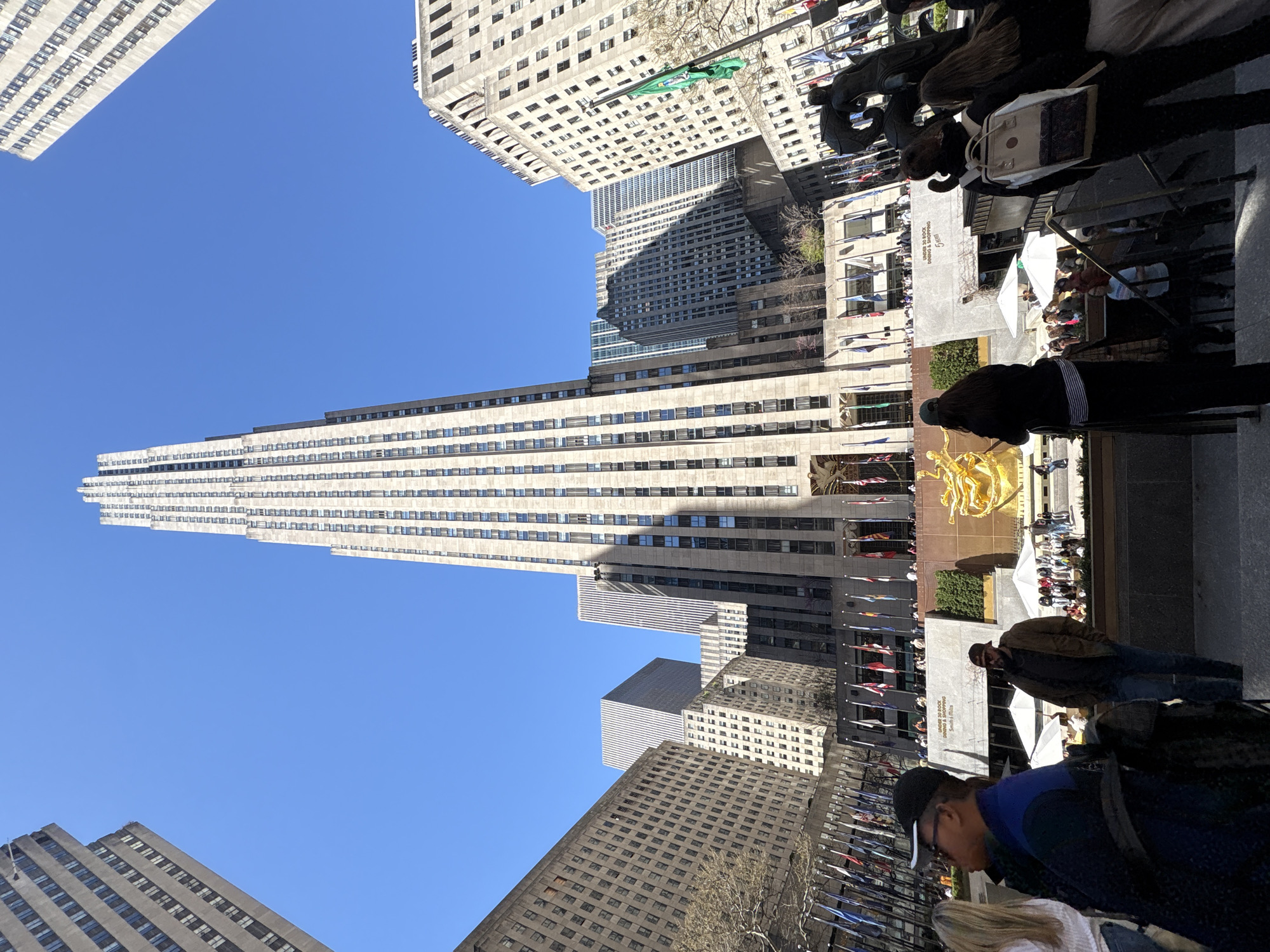 The image shows Rockefeller Center in New York City. Tall skyscrapers surround the plaza, and the sky is clear and blue. A golden statue stands prominently near an ice-skating rink. People are visible around the area, including some looking at the statue and others walking by. Flags are flying.