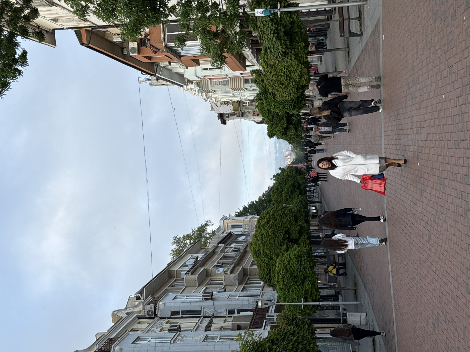 The image depicts a street scene in Varna, Bulgaria. The street is lined with buildings on either side, and trees are planted along the sidewalk. People are walking on the sidewalk, some heading towards the camera, others away. One woman, closer to the camera, is wearing a white coat and carrying a red bag. The sidewalk is paved with red bricks. Overhead, power lines stretch across the street between the buildings. The sky is overcast.