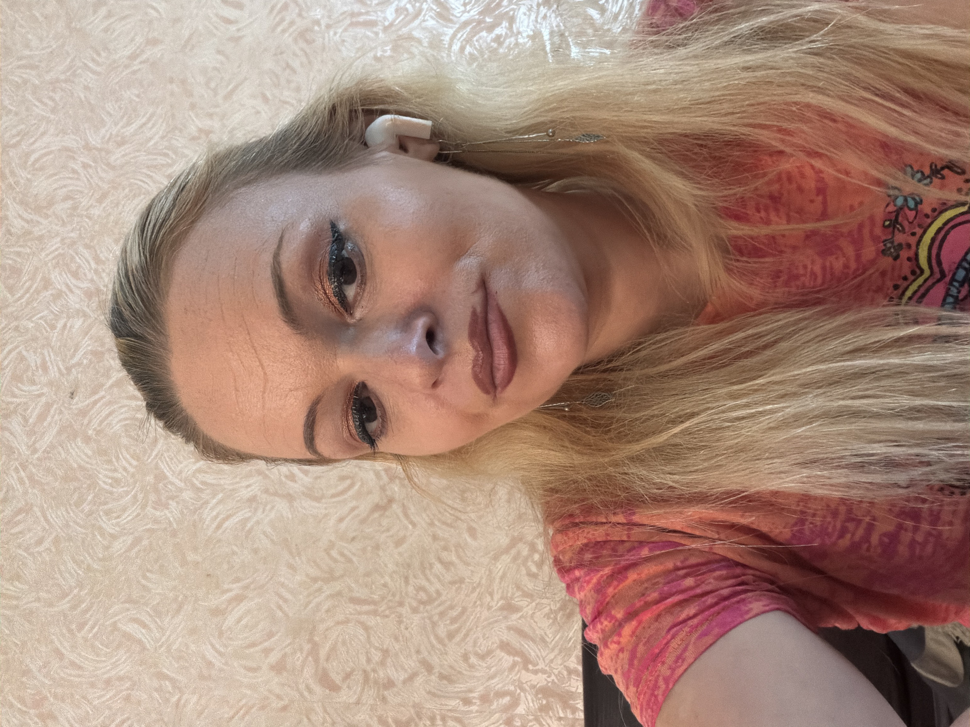 The image shows a close-up, head-and-shoulders selfie of a woman with blonde hair, wearing a pink and orange patterned t-shirt and white wireless earbuds. She has on makeup, including eyeshadow and lipstick. The background appears to be a textured, light-colored wallpaper. The image quality is moderate with some graininess. There are no discernible landmarks or contextual elements to place this image in Chernihiv, Ukraine.