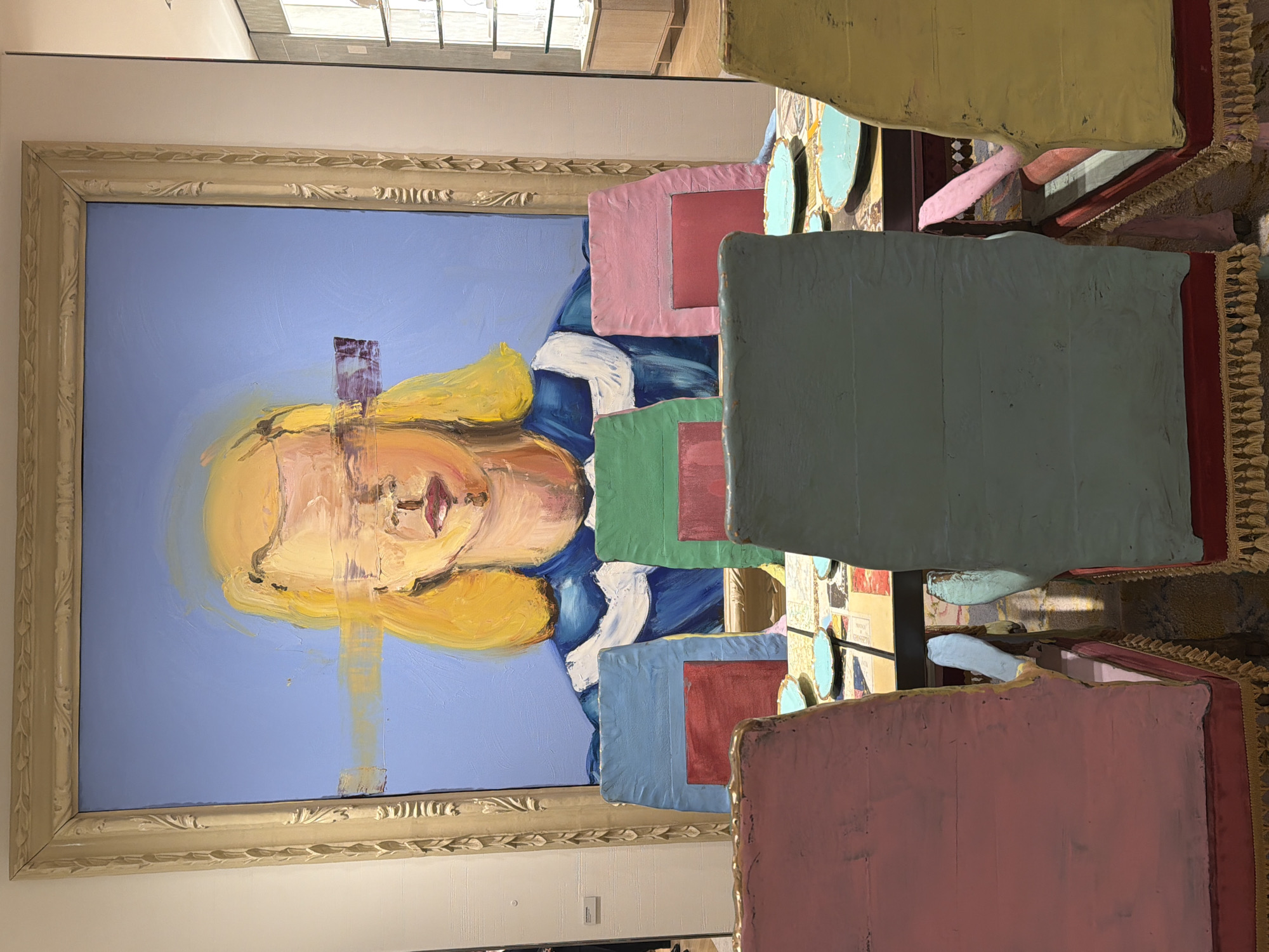 The image shows a portrait of a person with blonde hair in a blue and white dress, painted in a somewhat abstract style, hanging on a wall. The portrait is in an ornate gold frame. In the foreground, partially obscuring the portrait, are several square or rectangular painted surfaces in various colors (pink, blue, green, yellow) that appear to be parts of an art installation. Some of these surfaces have red squares within them. Other elements of the installation, including fringe or tassels and various textures, are visible. Based on the style of the painting and the overall art installation, it is likely located in an art gallery or museum. Without additional context, it's impossible to determine the precise location in New York City, USA.