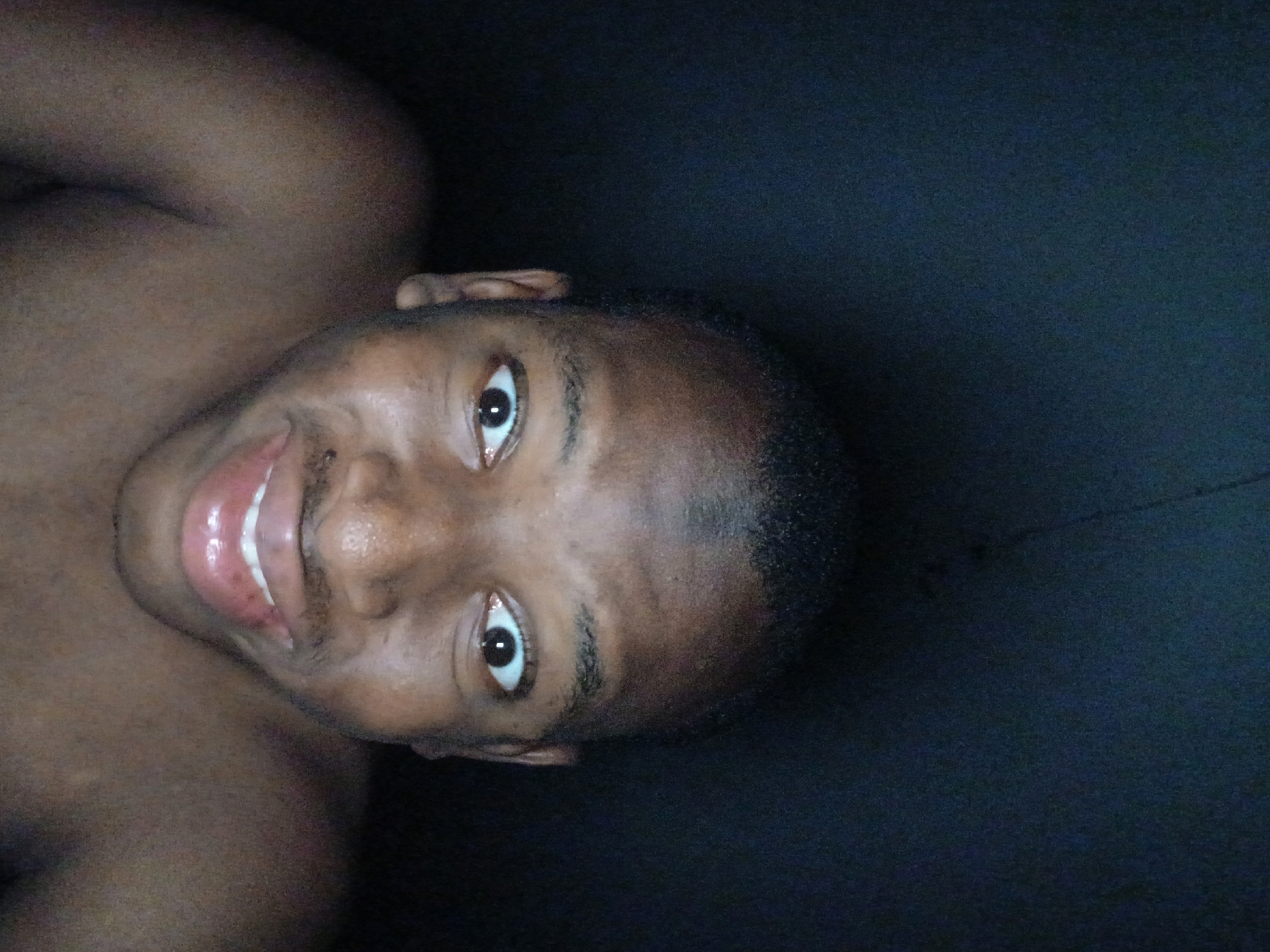 A medium, close-up shot of a dark-skinned, Black male’s face with dark hair, appearing to be clean shaven. The subject is smiling, showing his teeth. The background is predominantly a dark color. The image may have been taken indoors in a low-lit environment. There is no information to indicate the location as Lagos, Nigeria.